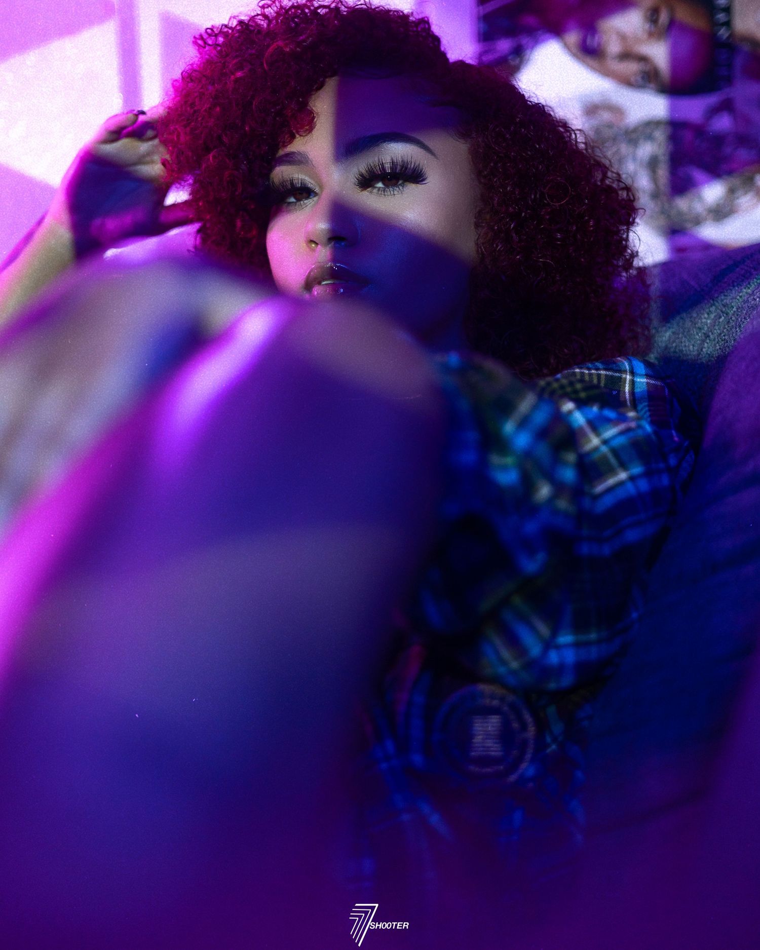 Portrait photo in purple lighting showing a person with curly burgundy hair wearing dramatic eye makeup and a plaid shirt.