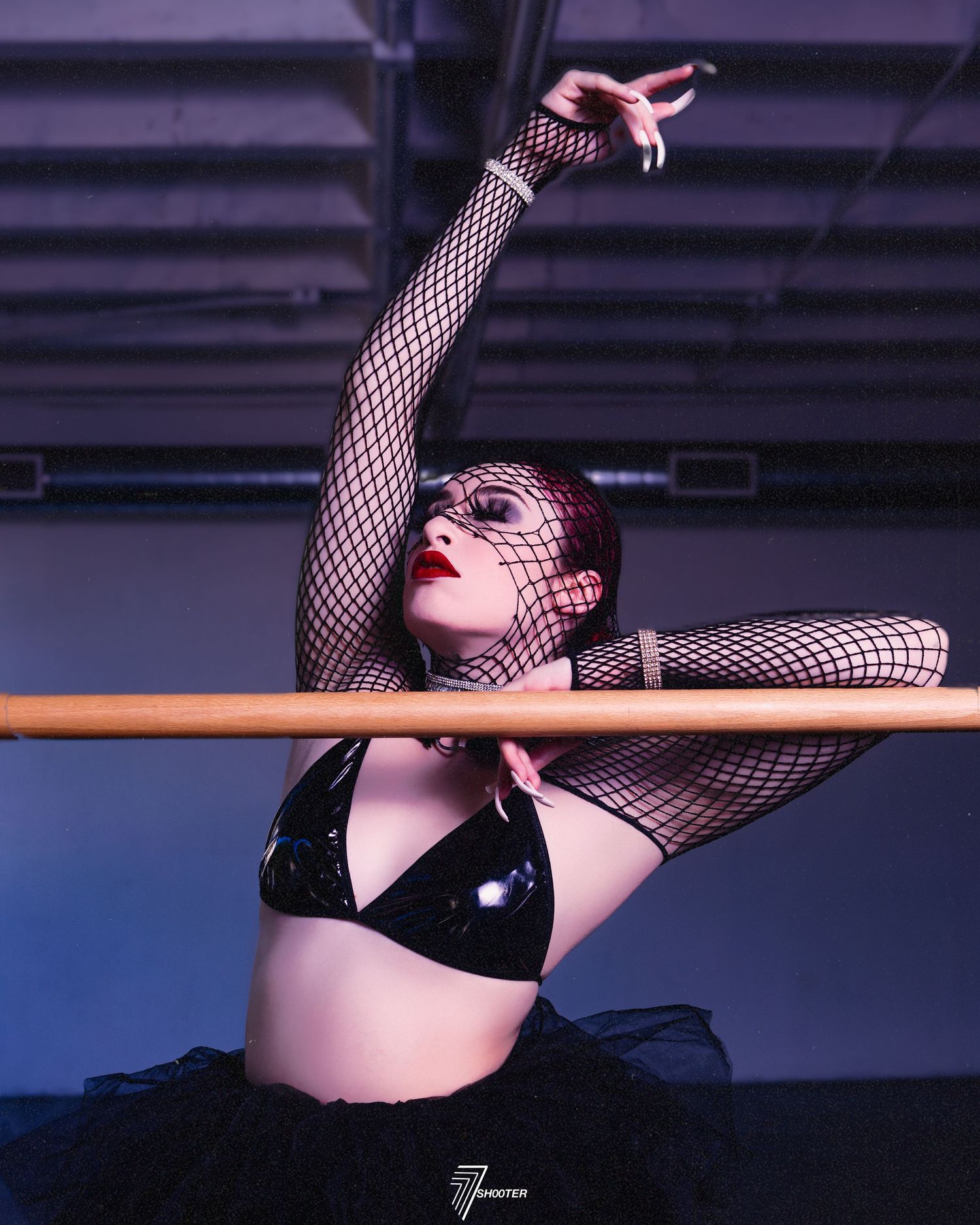 Performer in black bikini top and fishnet attire strikes dramatic pose on ballet barre in moody purple lighting.
