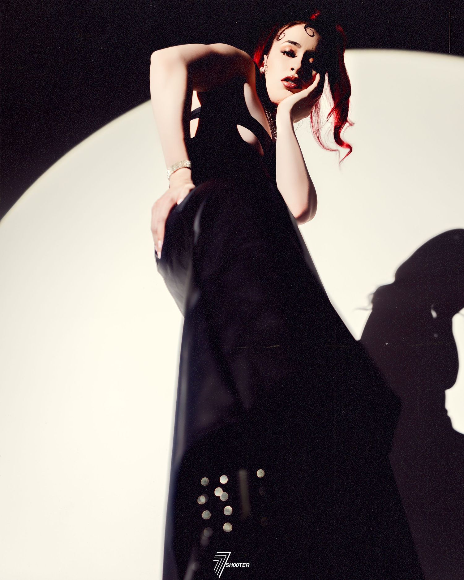 Dramatic fashion photo of a figure in a long black dress posing against a stark white circular backdrop with cast shadows.