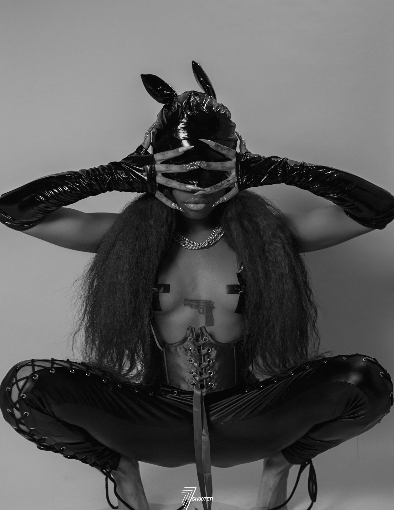 Black and white artistic portrait showing figure in latex mask and gloves with long dark hair in dramatic pose.