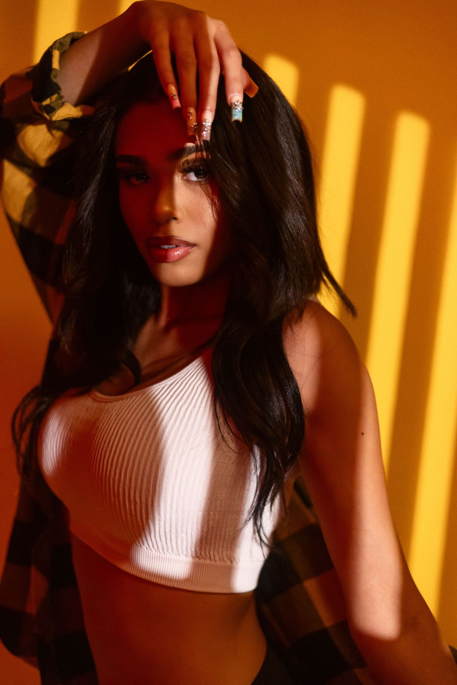 Studio portrait with dramatic orange lighting of a model in a white tank top against a wall with vertical shadow patterns.