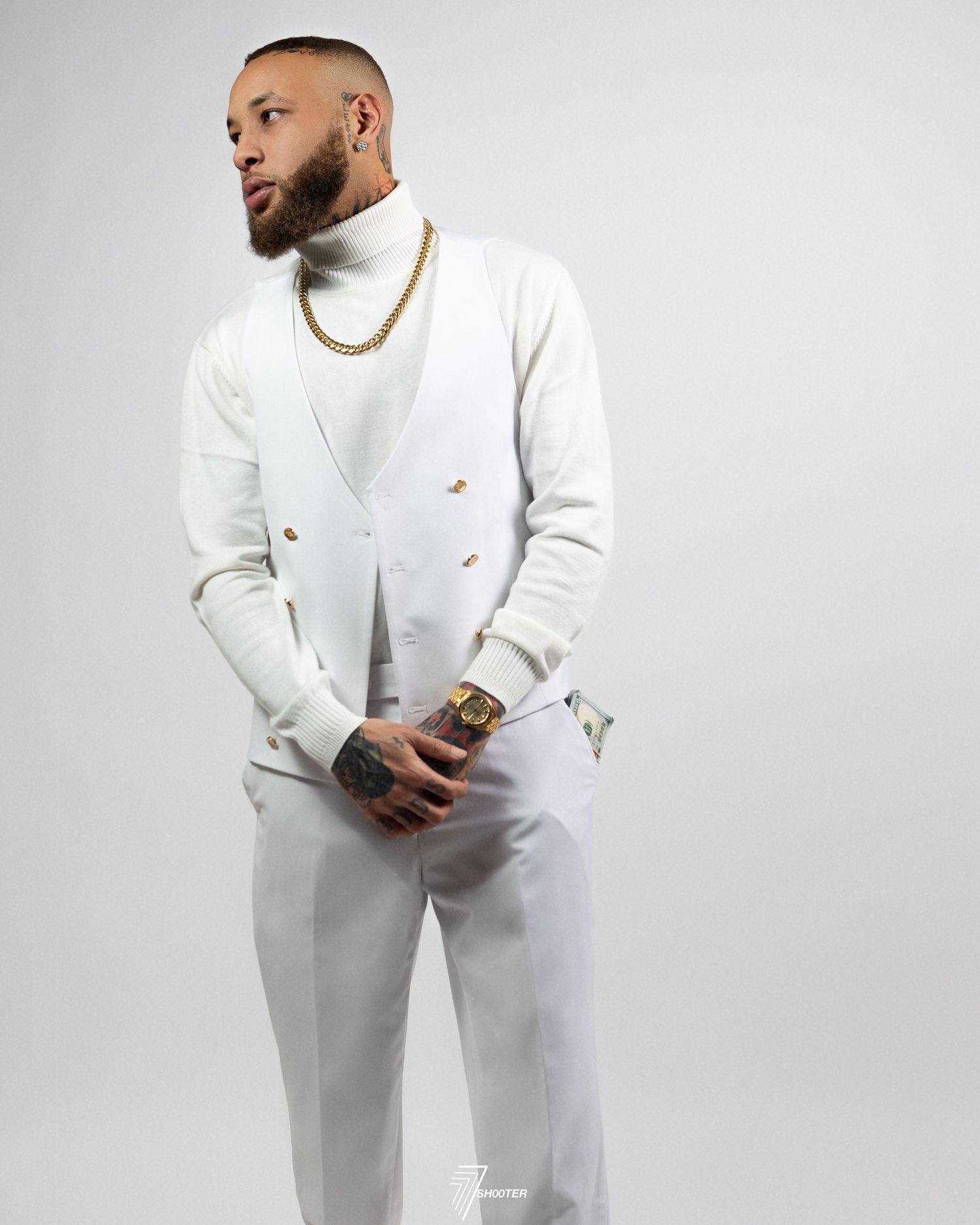 A figure wearing an all-white suit with gold chain necklace poses against a light grey background in a studio setting.