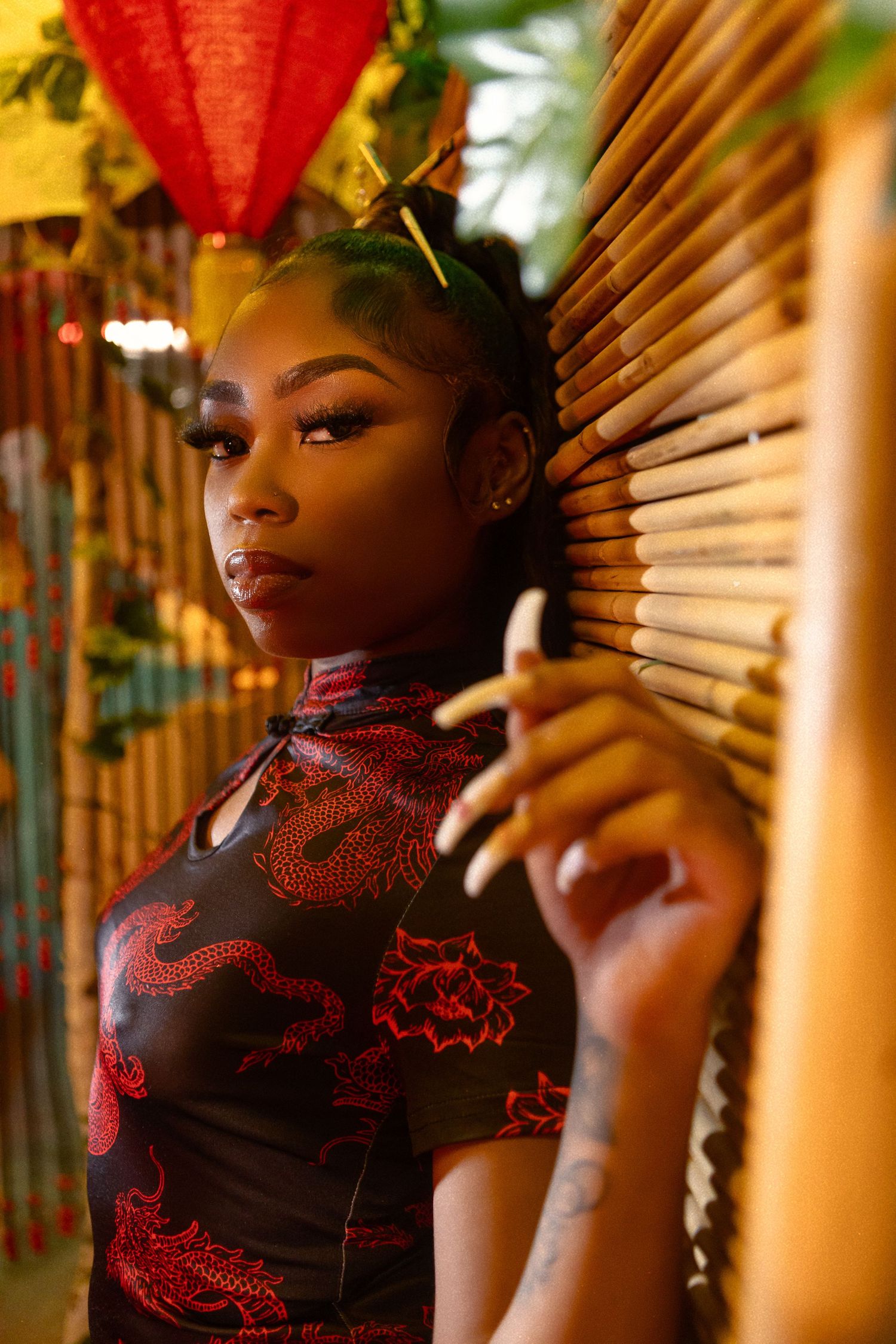 A person in a black and red qipao dress poses beside bamboo blinds in an Asian-themed setting with red lantern decor.