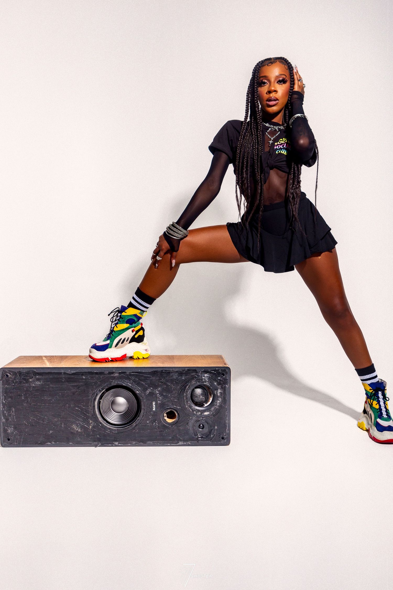 A model poses on a speaker wearing a black sheer outfit, colorful sneakers, and athletic socks in a dynamic fashion shot.