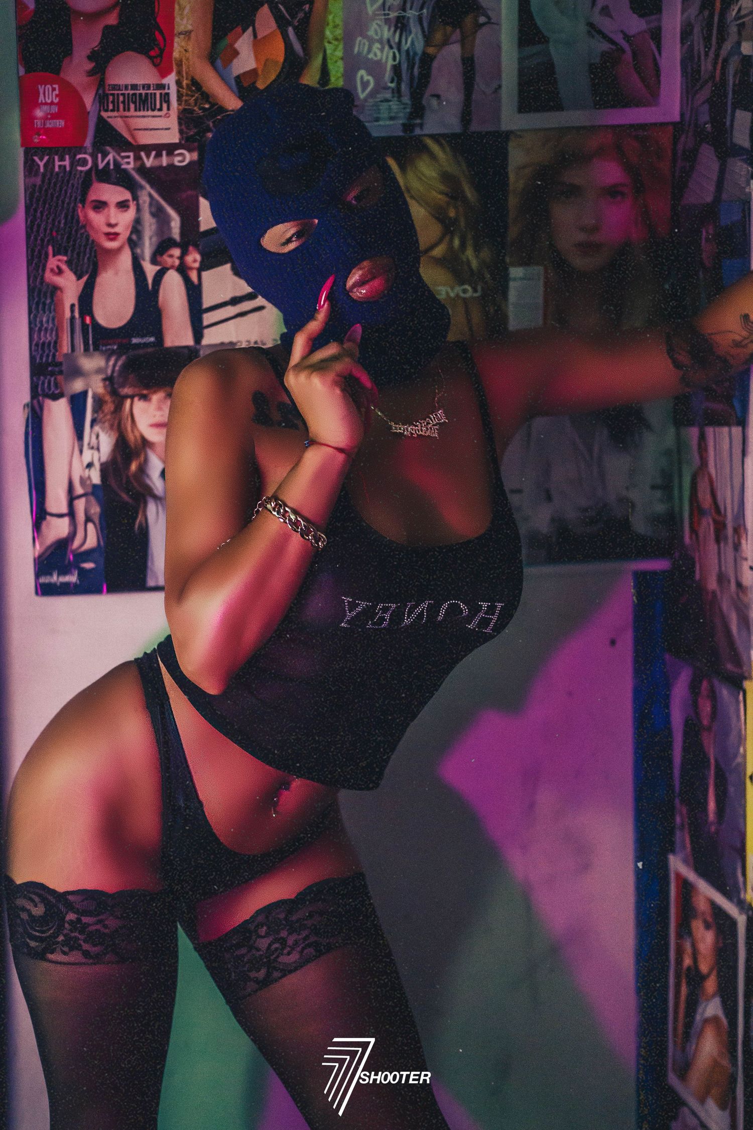 A person wearing a black balaclava mask poses seductively in lingerie against a colorful backdrop with posters.