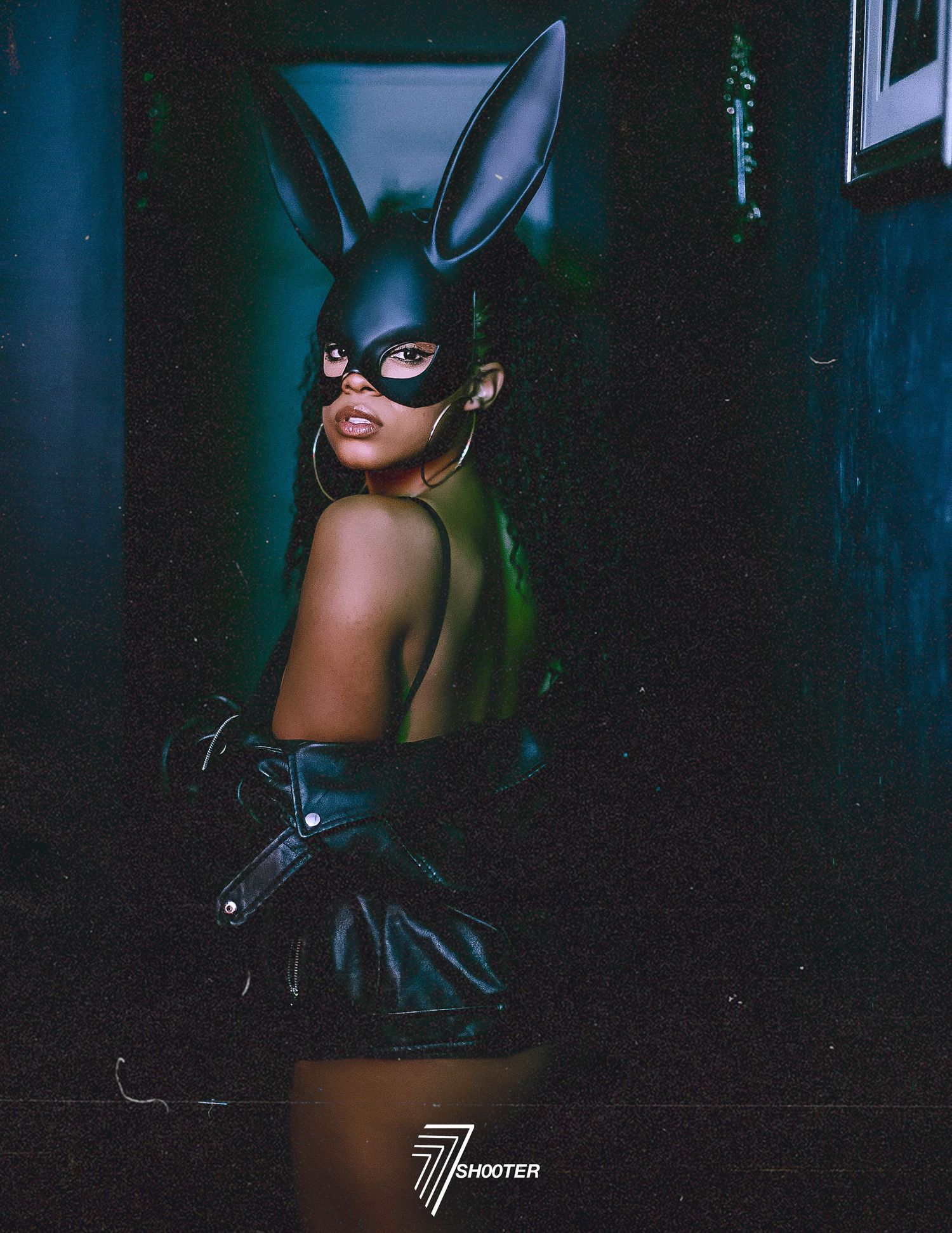 A figure in a black bunny mask and dark attire poses against a moody blue-lit backdrop in an artistic photoshoot.