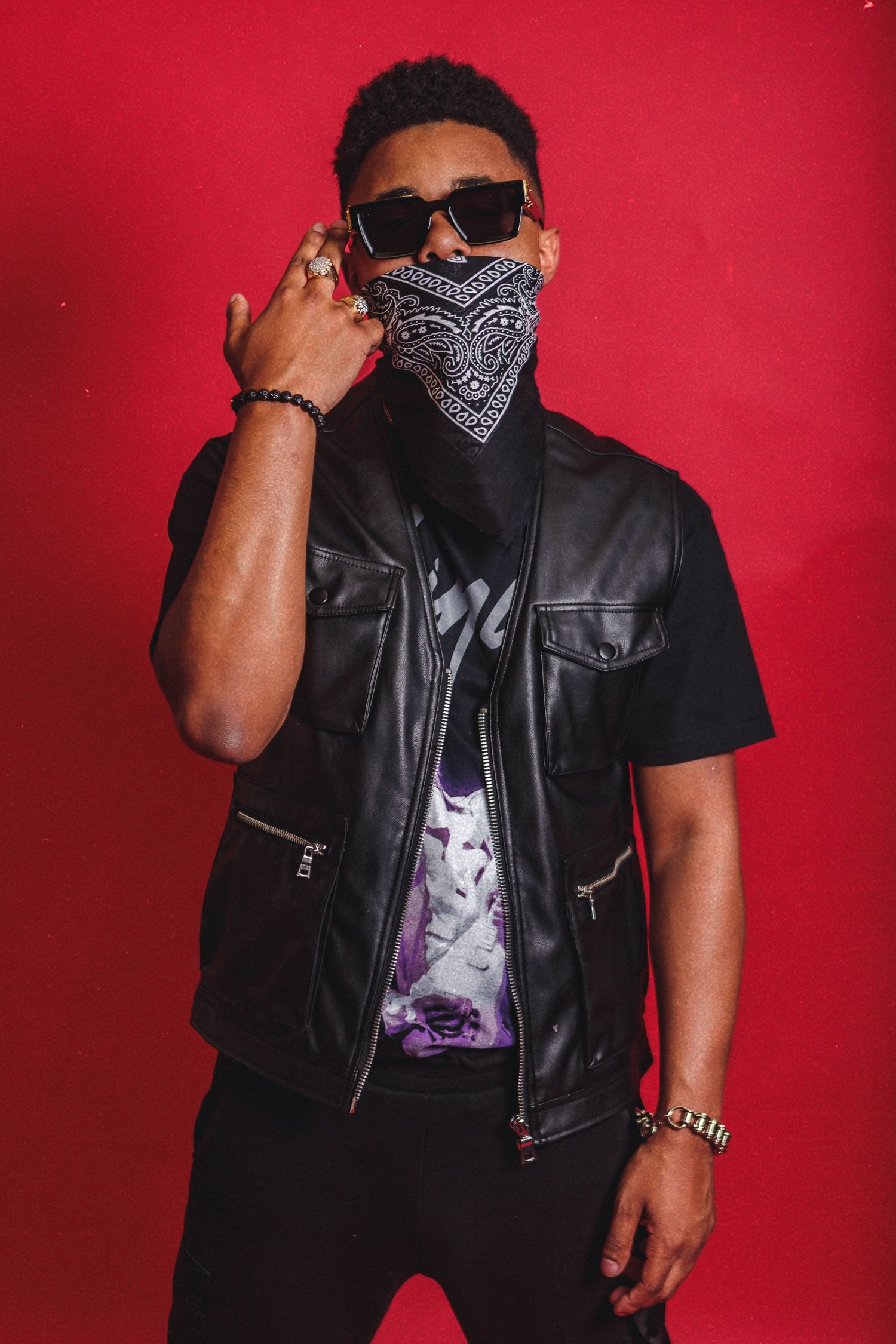 Person in black leather vest and bandana mask poses against red background wearing sunglasses and casual streetwear.
