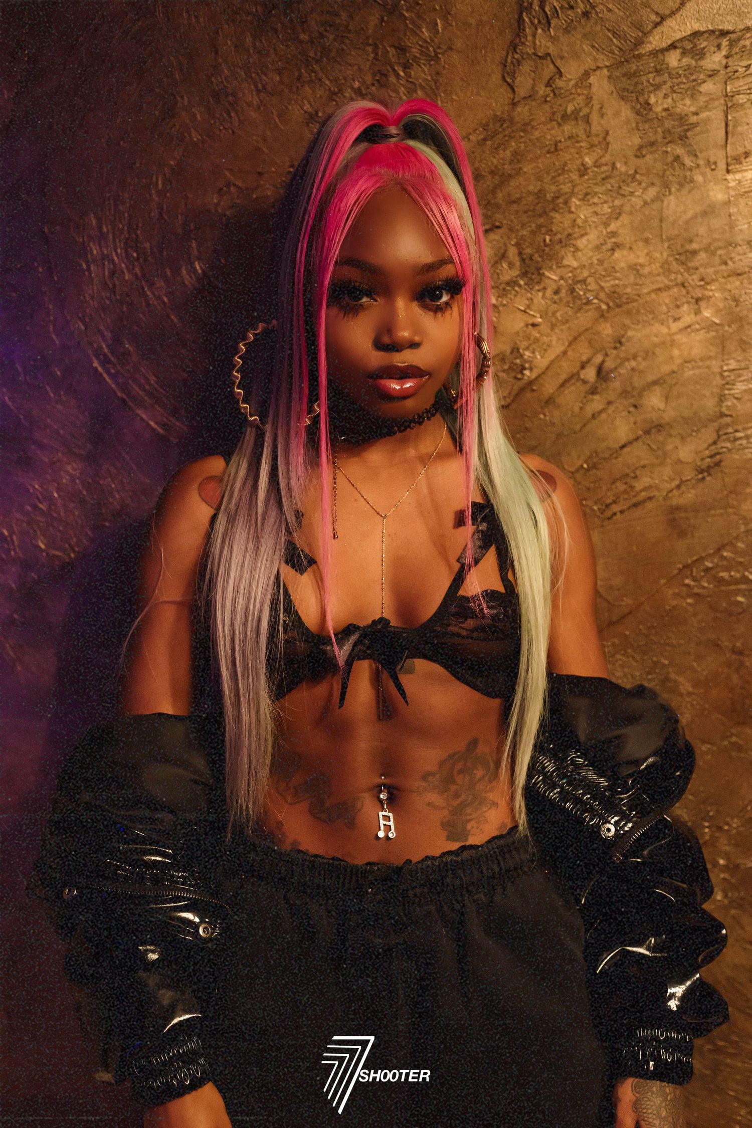 A model in a black crop top and shiny pants poses against a textured brown wall, wearing vibrant pink and silver hair.