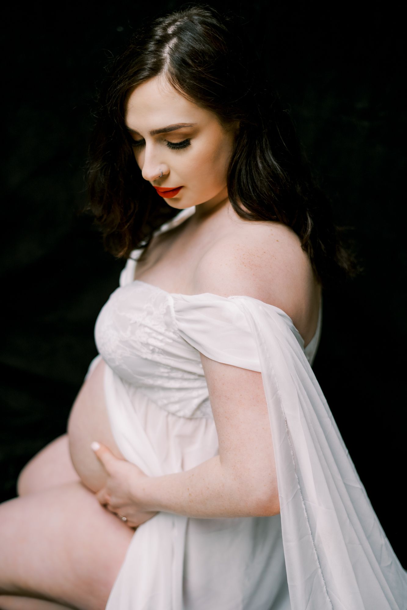 A maternity portrait in white flowing fabric against a dark background shows an expectant mother looking down gracefully.