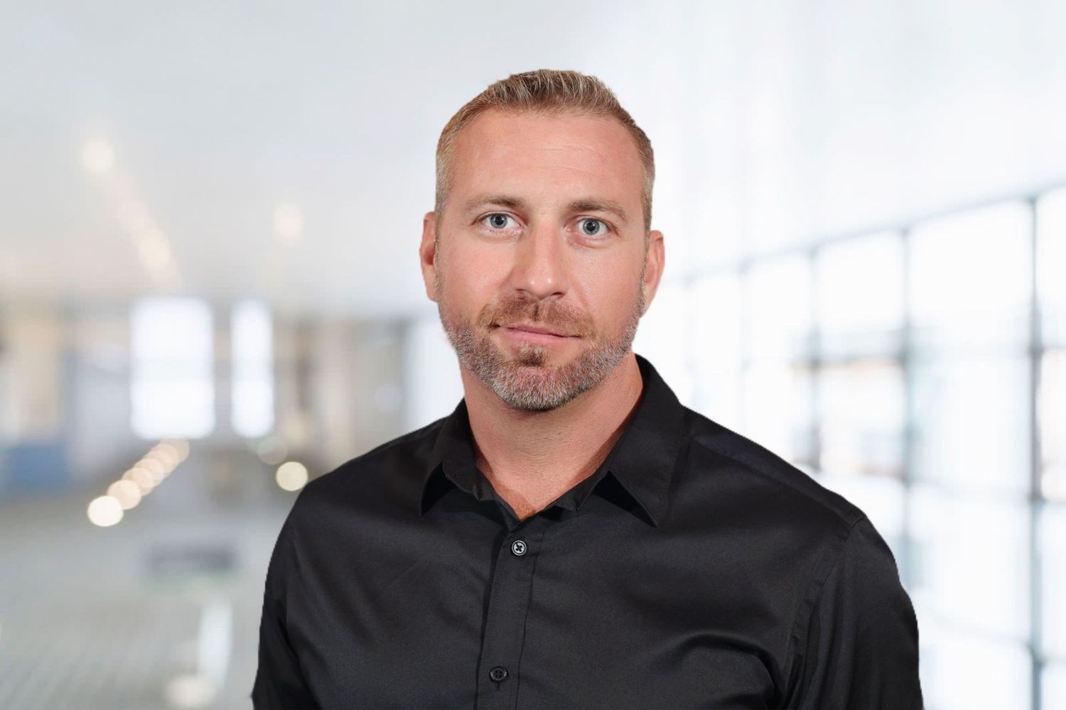 Professional man in black shirt with short blonde hair posing in modern office setting.