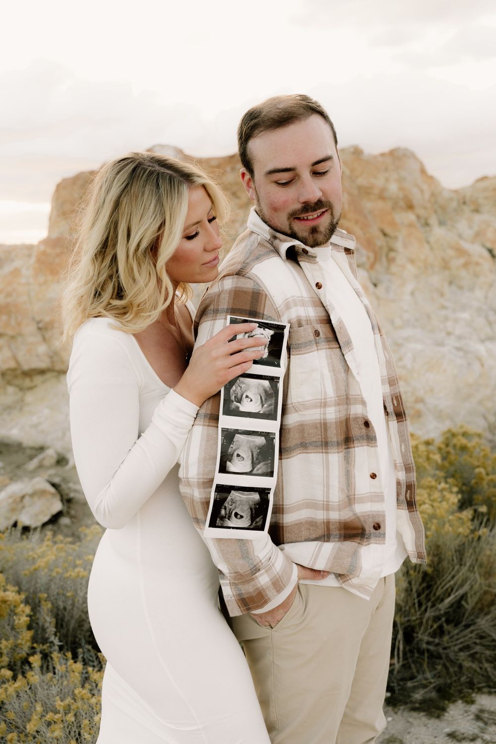 Couple holding ultrasound photos during pregnancy announcement photoshoot near rocky cliffs.