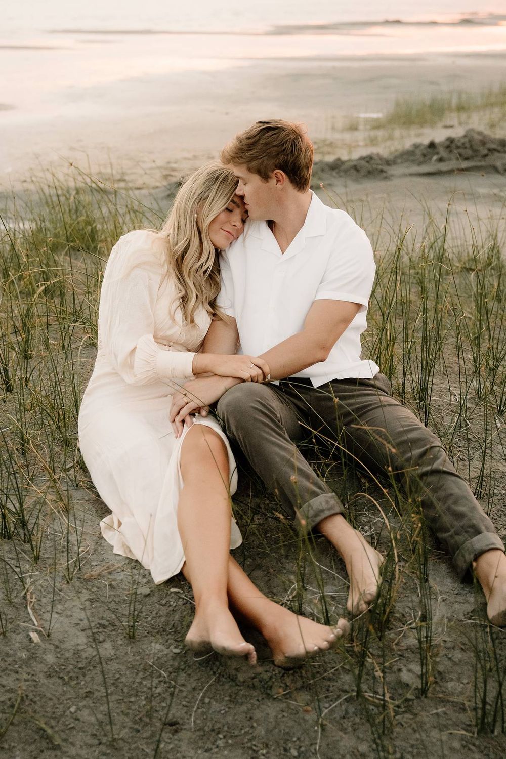 A romantic couple sits intimately on a beach at sunset in casual attire.