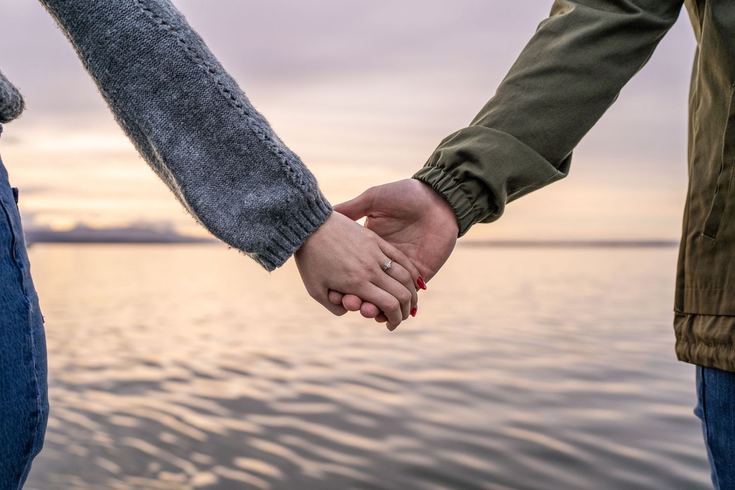Close-up of couple holding hands against sunset waterfront backdrop.