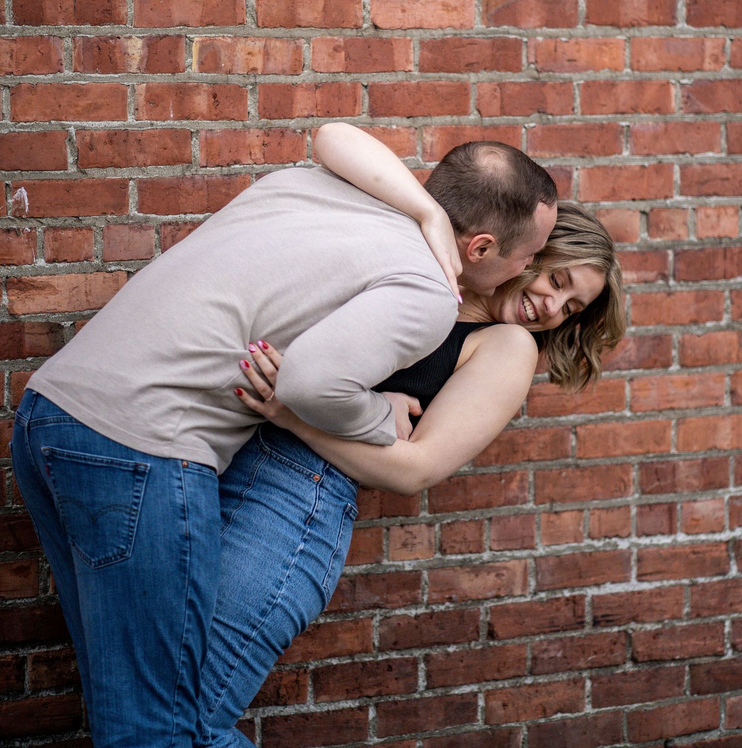 Playful couple embracing against red brick wall during engagement photo session.