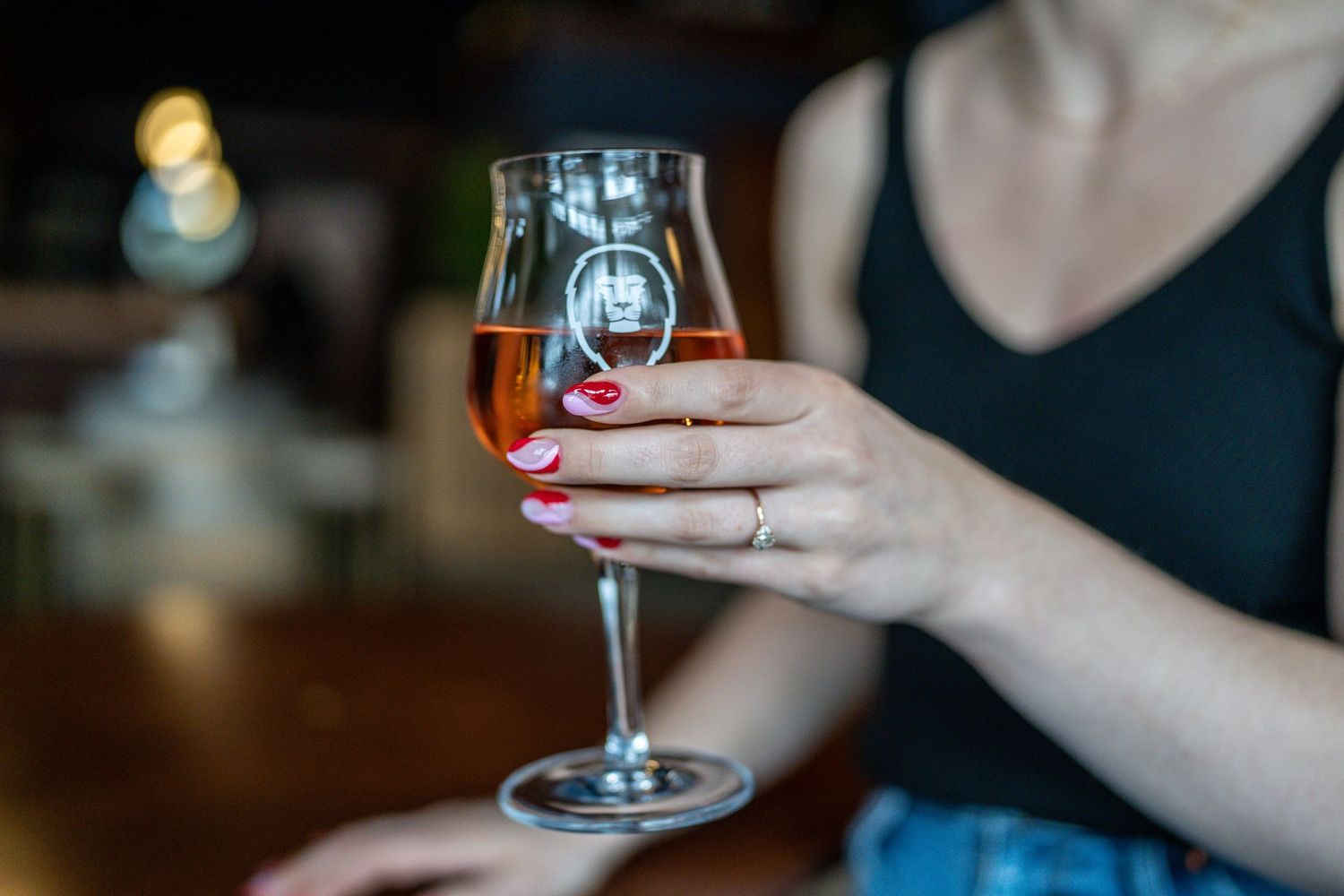 Hand holding a stemmed glass of amber beer or wine with red nail polish in a dimly lit setting.