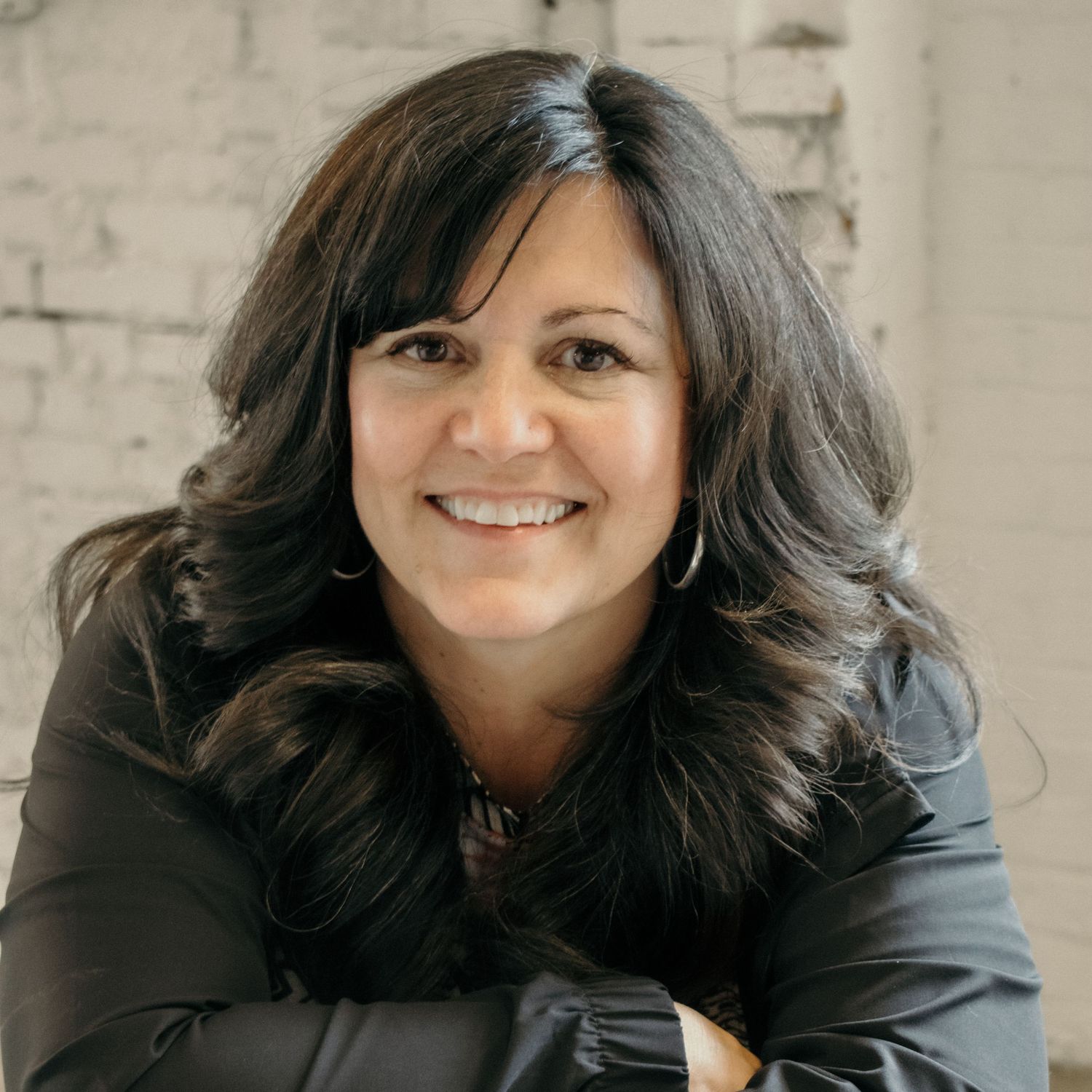 Cheerful woman with dark curly hair wearing black against white brick wall.