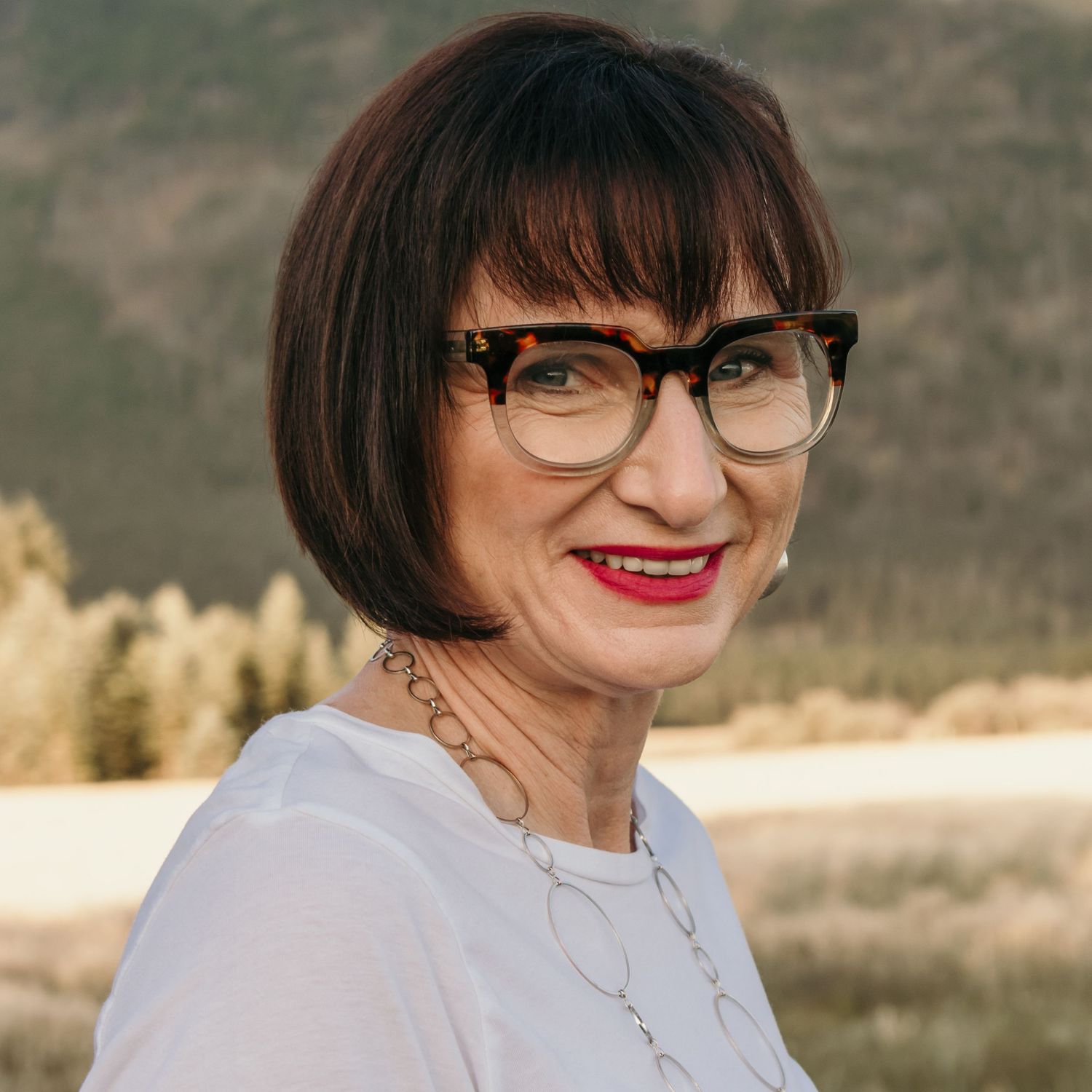 Portrait of woman with short dark hair wearing glasses and white top against mountain backdrop.
