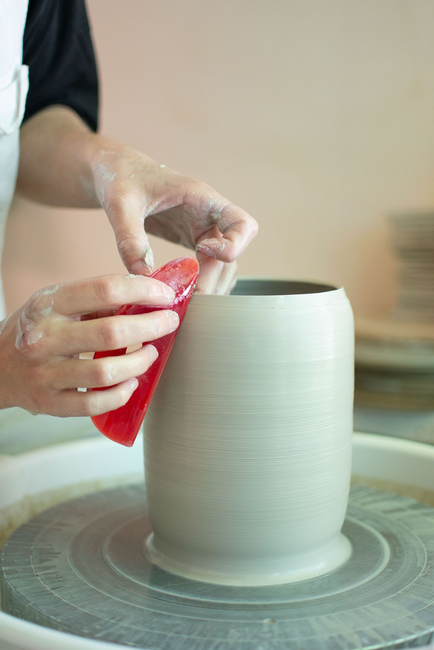 Hands smoothing a clay vessel on a pottery wheel with a red tool.