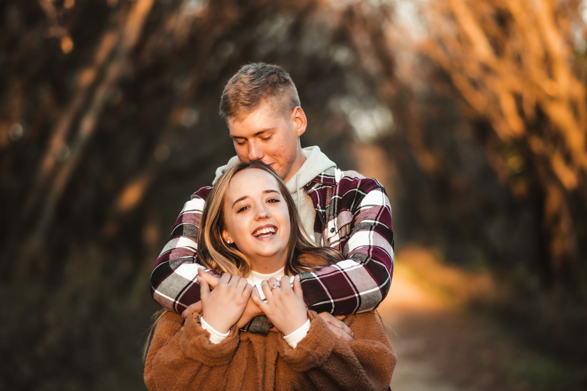Playful couple embracing and laughing on autumn path with golden sunset behind them.