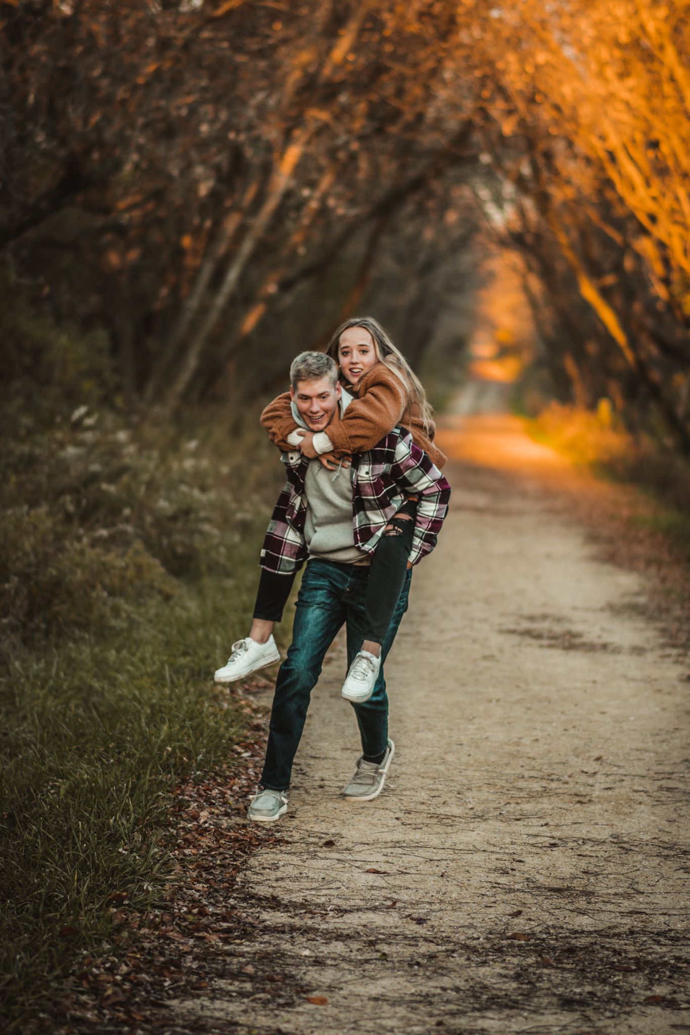 Two people playfully piggyback riding on a dirt path surrounded by trees with golden autumn foliage.