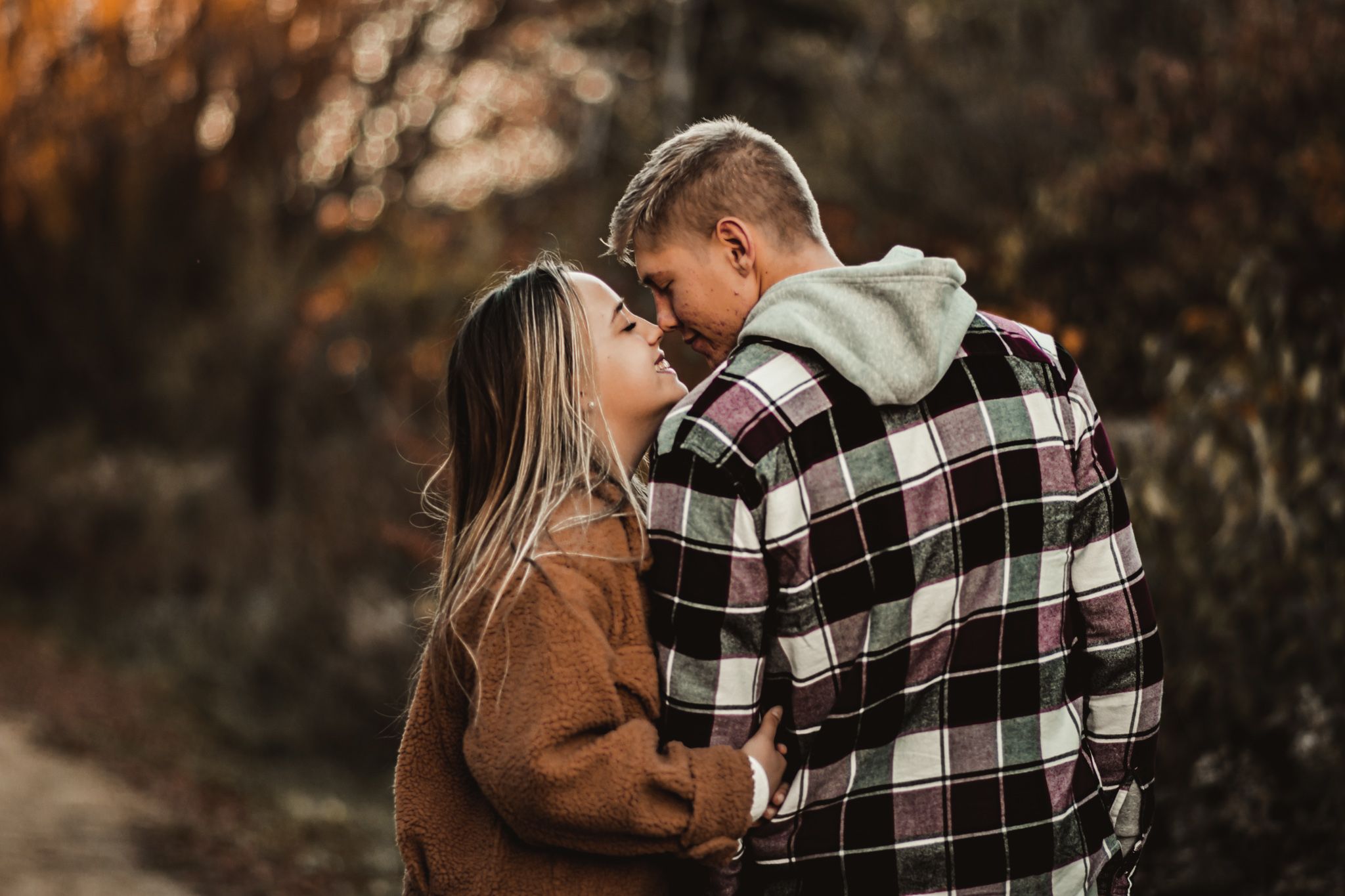 A romantic couple in fall fashion embraces on a nature trail with warm autumn colors in the background.