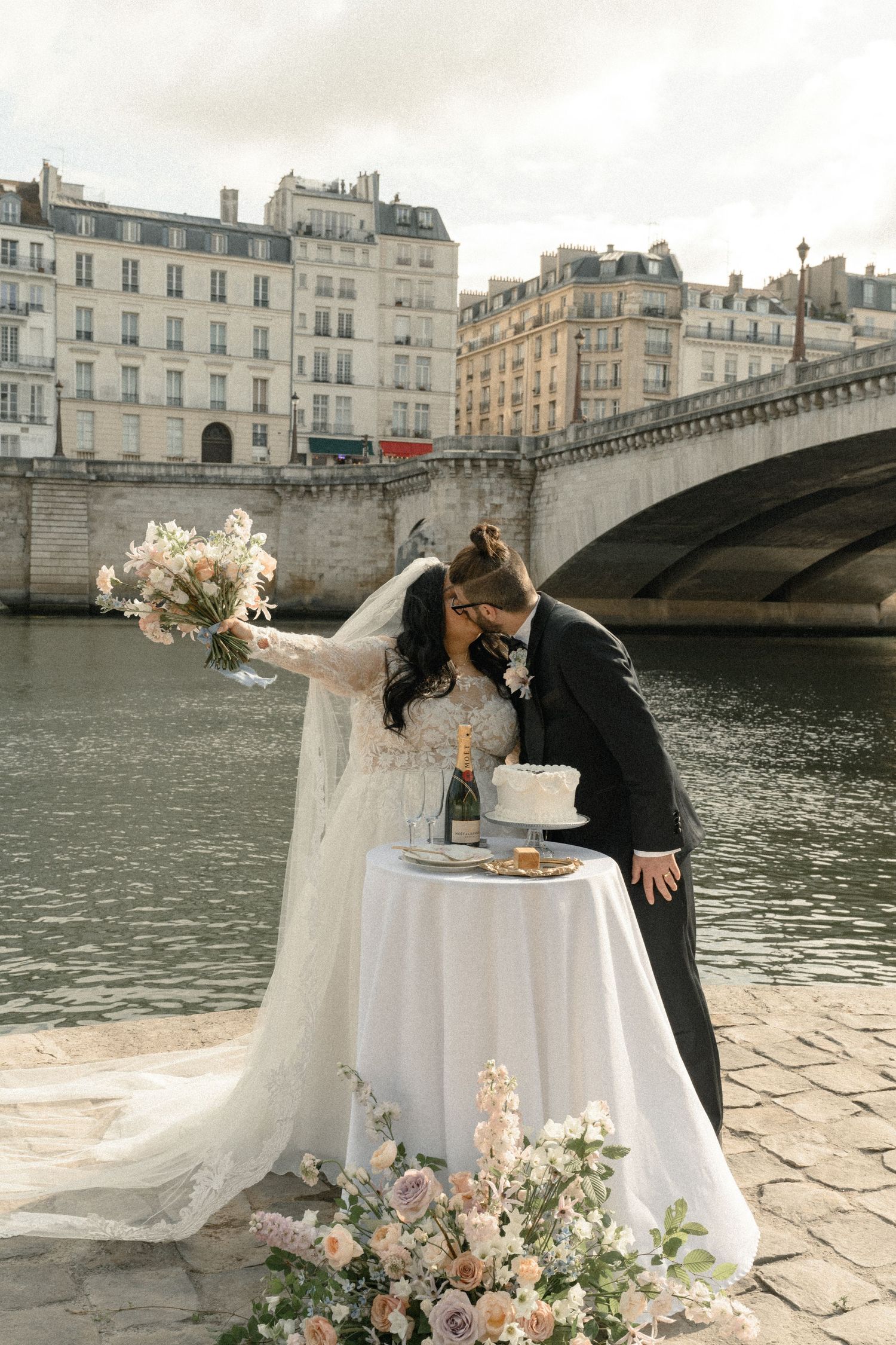 Newlyweds sharing an intimate moment by the Seine River with a decorated table and floral arrangements.