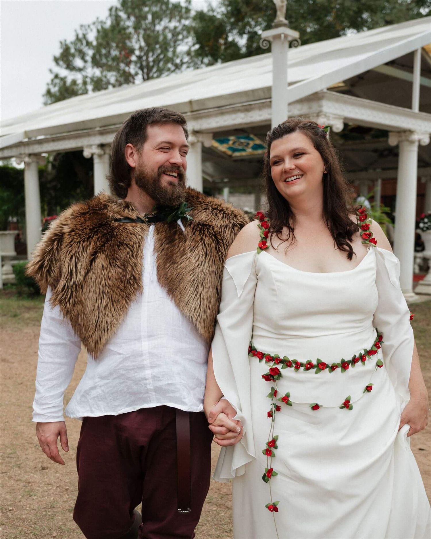 Renaissance wedding couple in fur and floral garments pose together on dirt path under white structure.