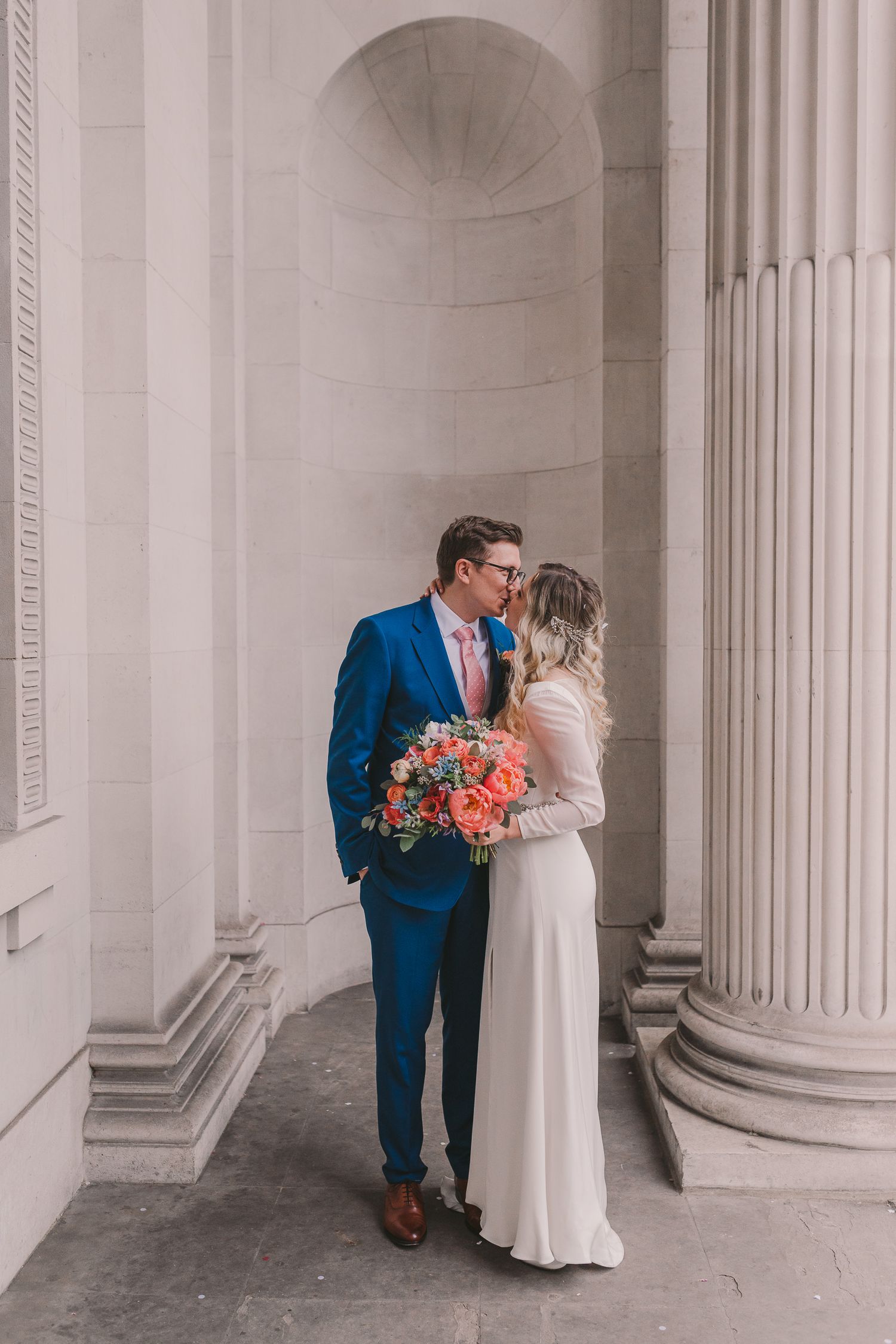 Couple shares intimate moment in elegant archway with coral bouquet and classic formal attire.