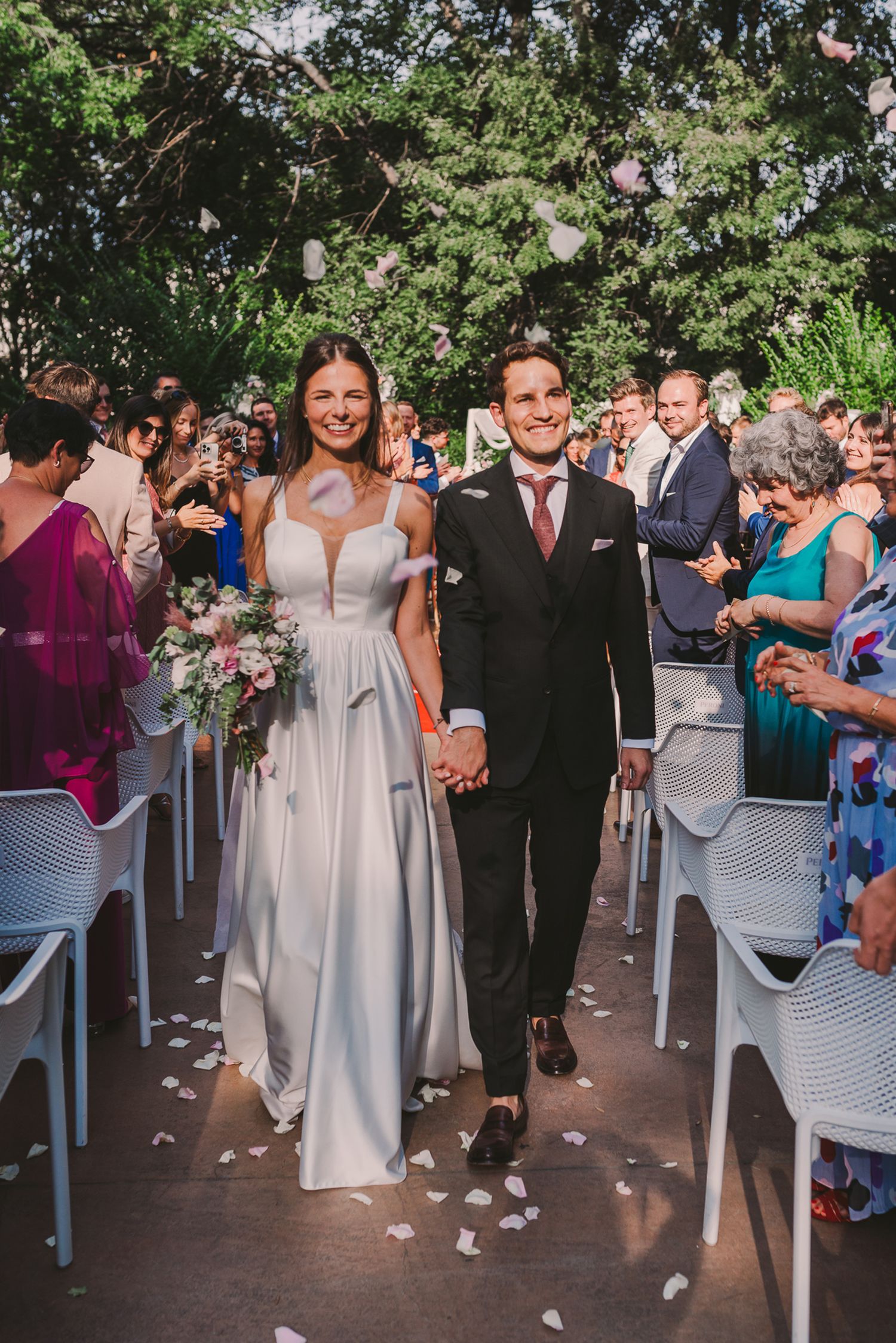 Wedding party walking down an outdoor aisle while guests throw flower petals.
