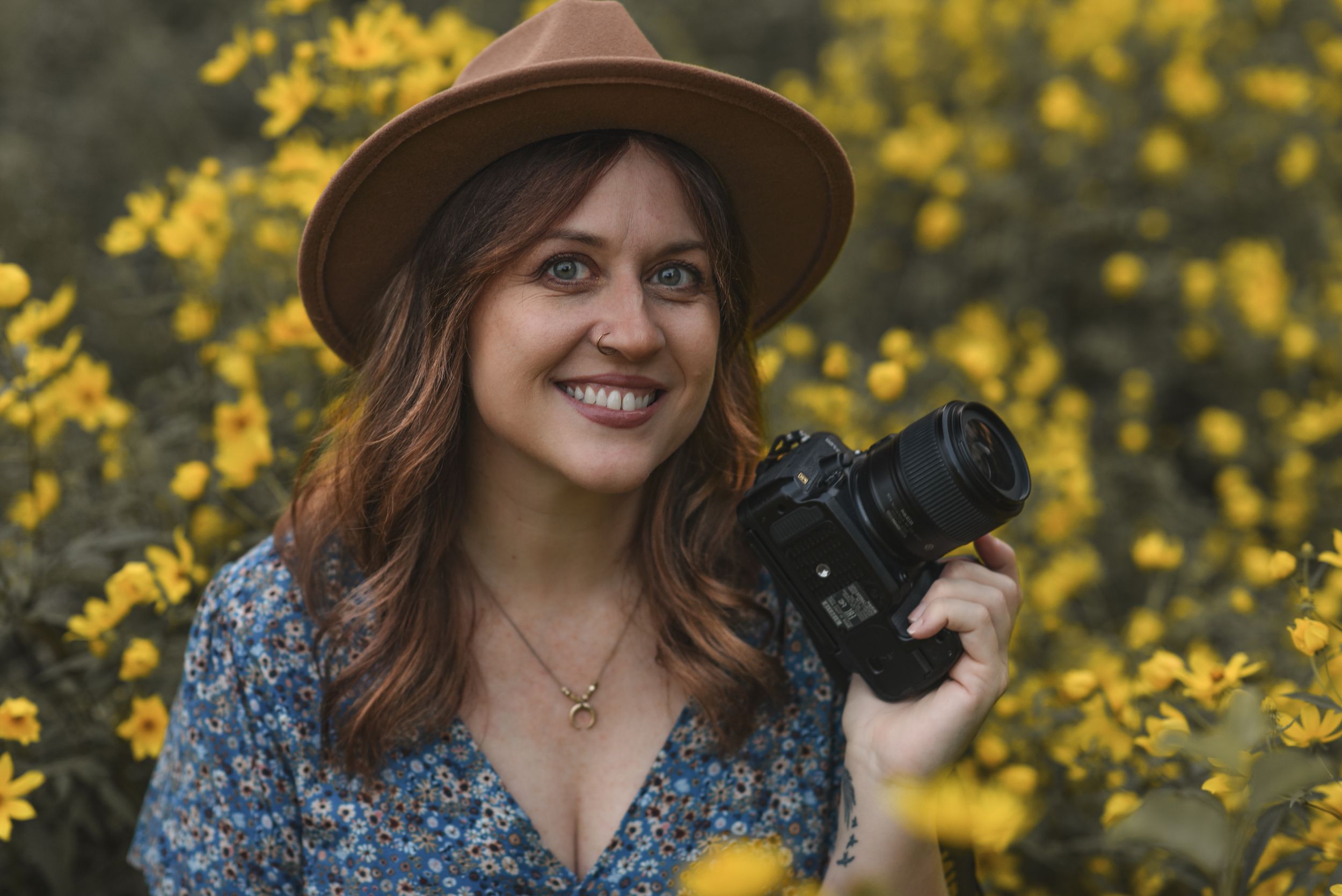 Photographer with brown hat and floral dress holds camera against backdrop of yellow flowers.