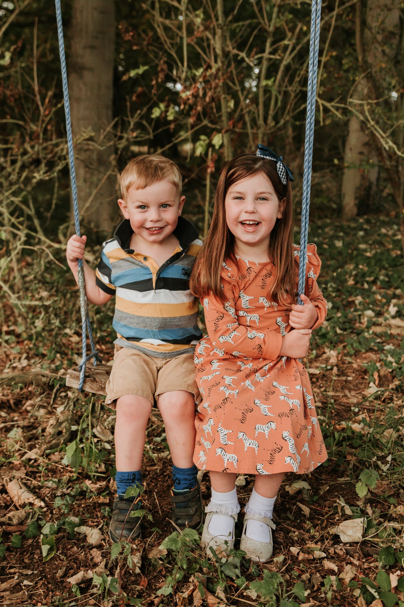 Two children sit together on a wooden swing in an autumn garden setting.