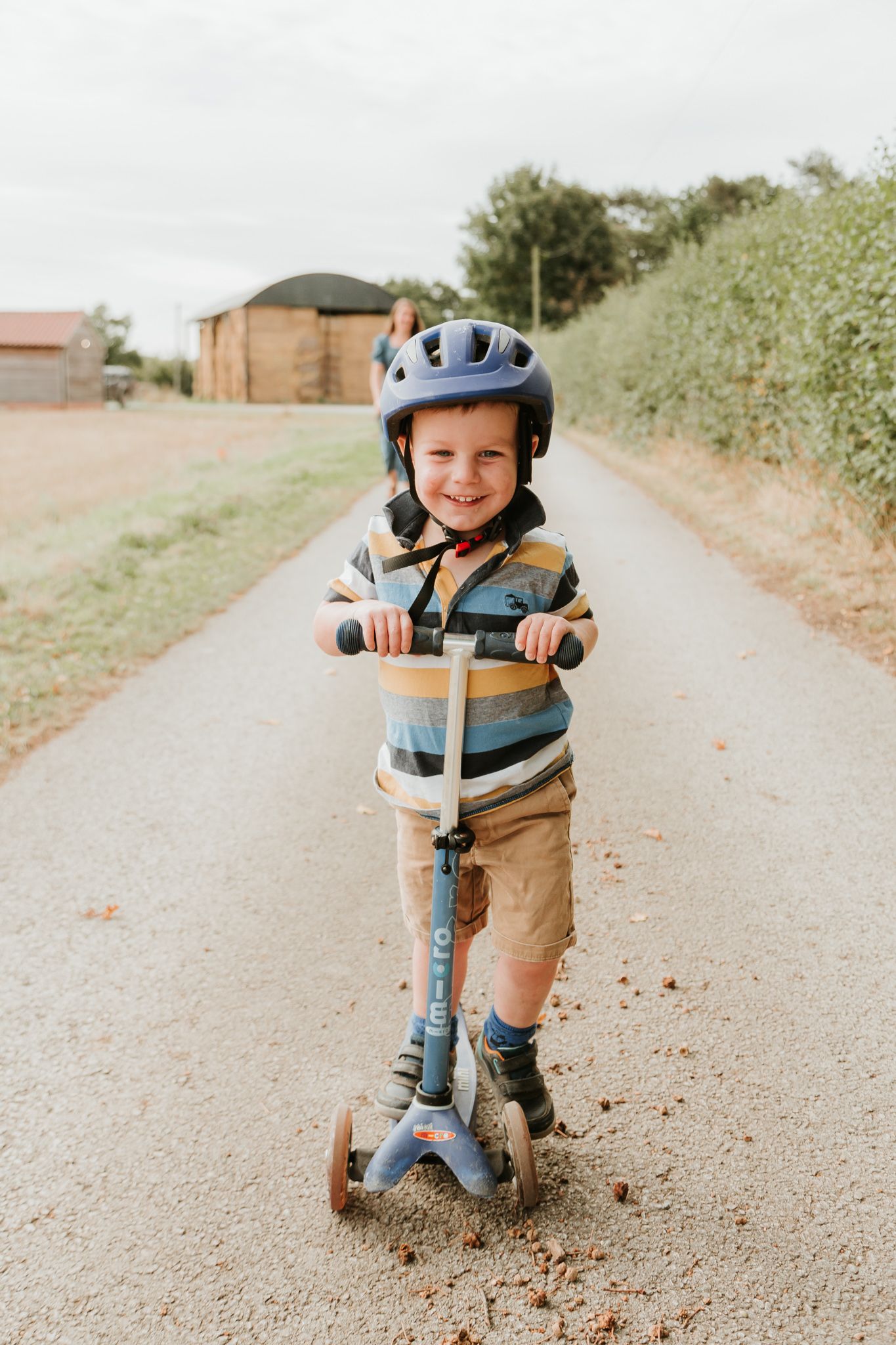 A child rides a blue scooter on a path while wearing safety gear and a striped shirt.