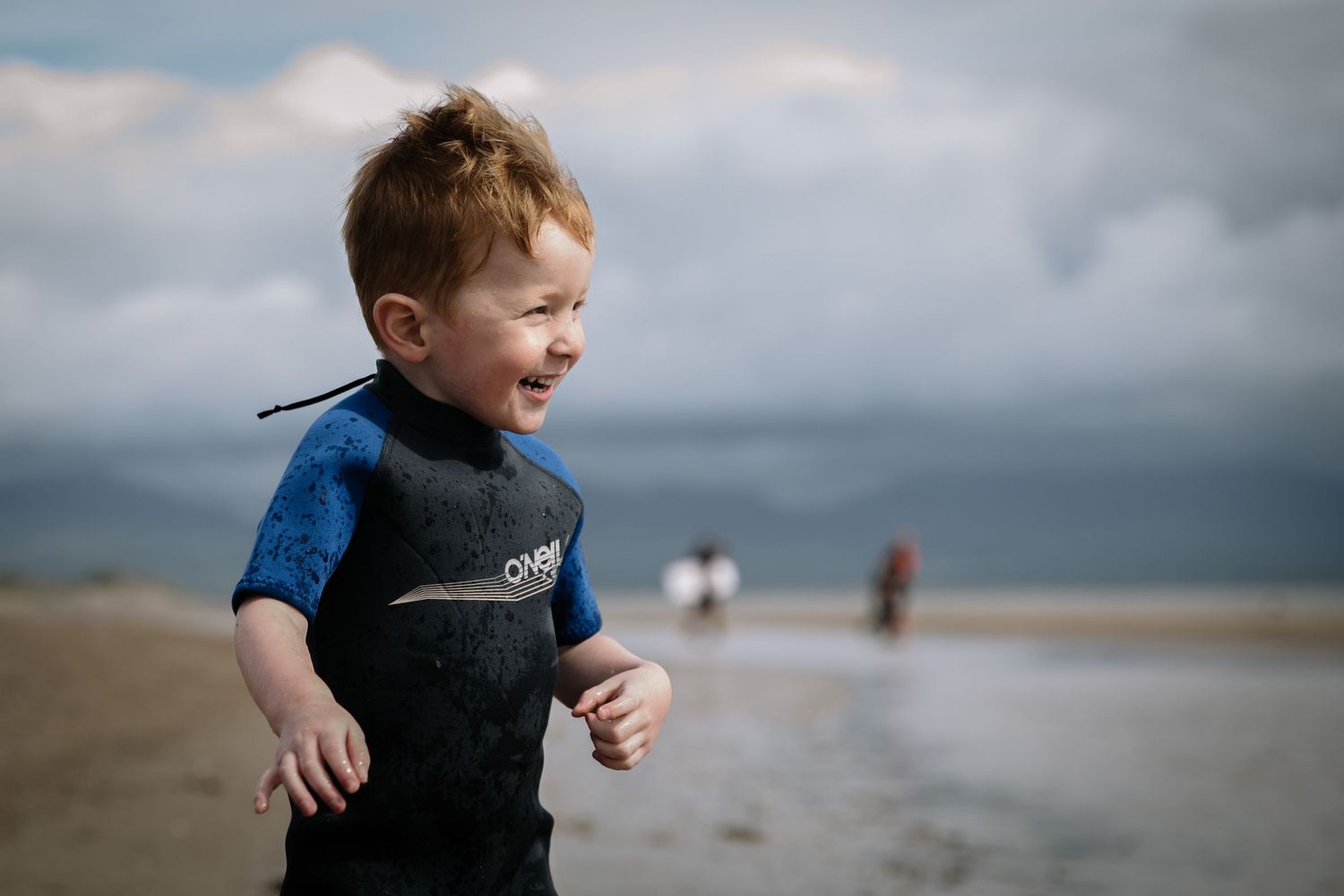 Child in a blue wetsuit laughing on a cloudy beach.