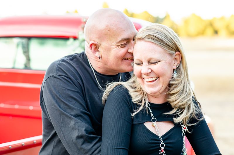 Husband nuzzles his wife as she giggles while they sit on a vintage red truck in Colorado Springs, Colorado.
