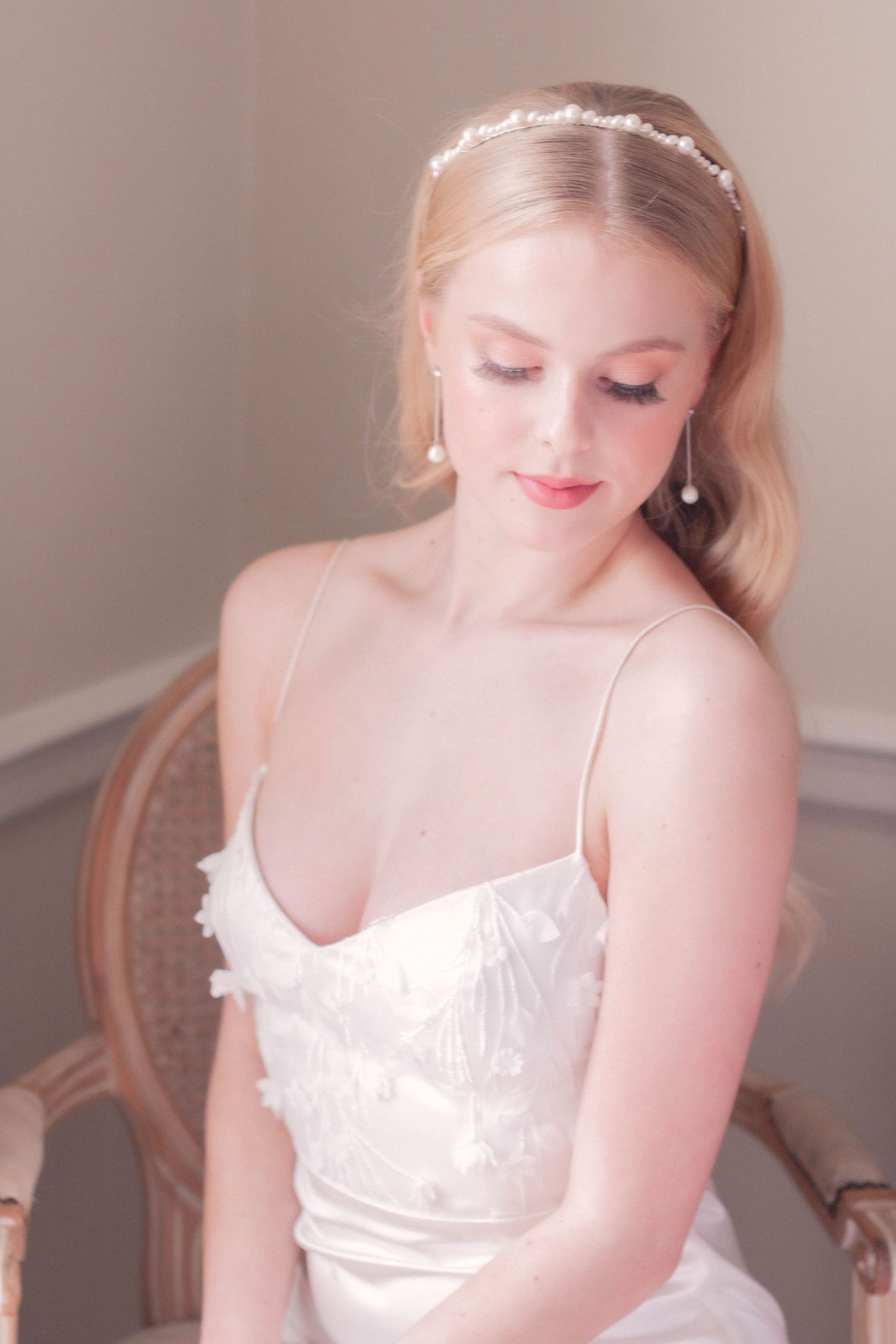 Bride in white satin dress with delicate lace details and pearl headpiece looking down with soft makeup.