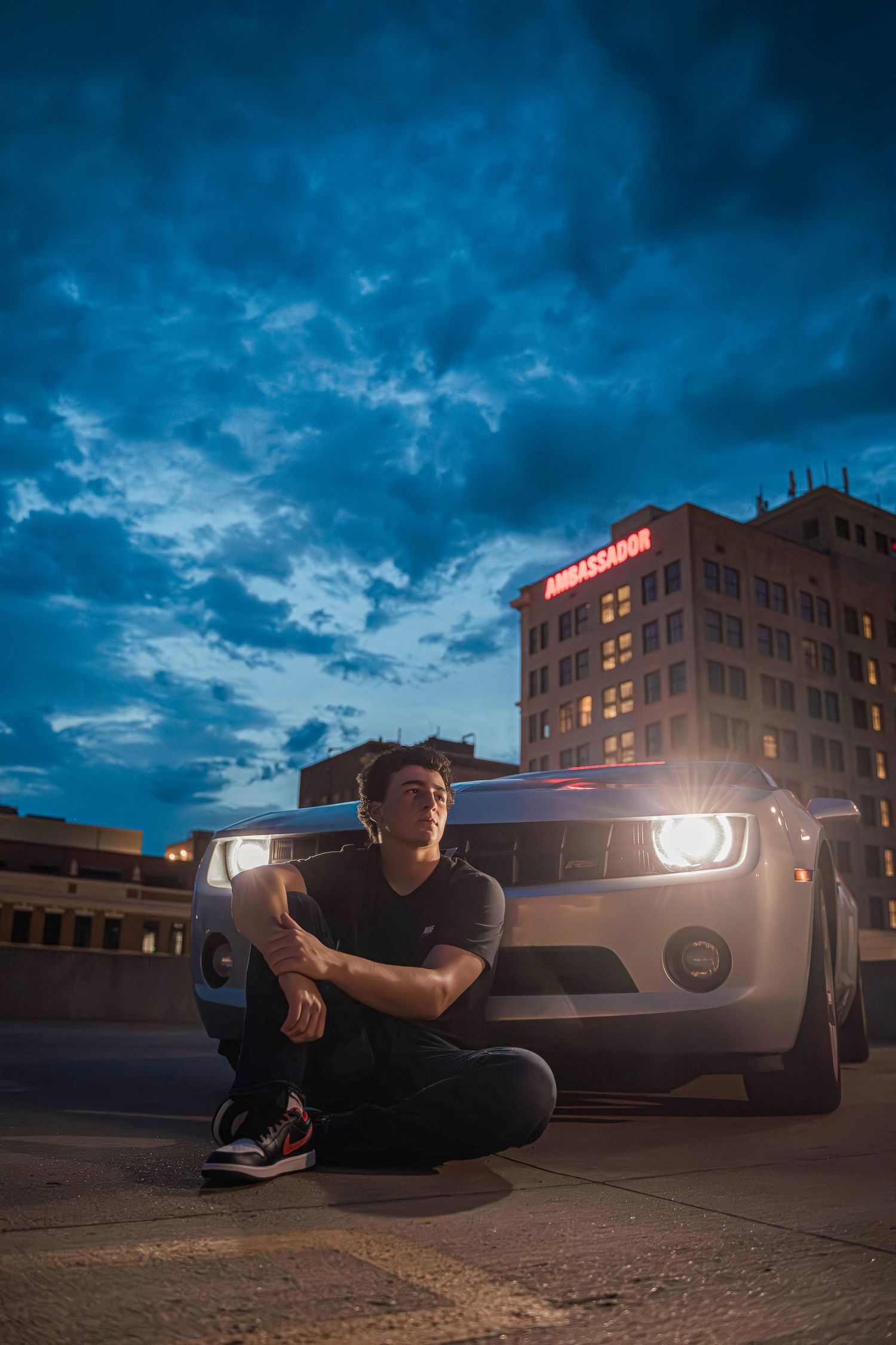 Urban nighttime photo with person seated in front of car with headlights on against dramatic blue cloudy sky.