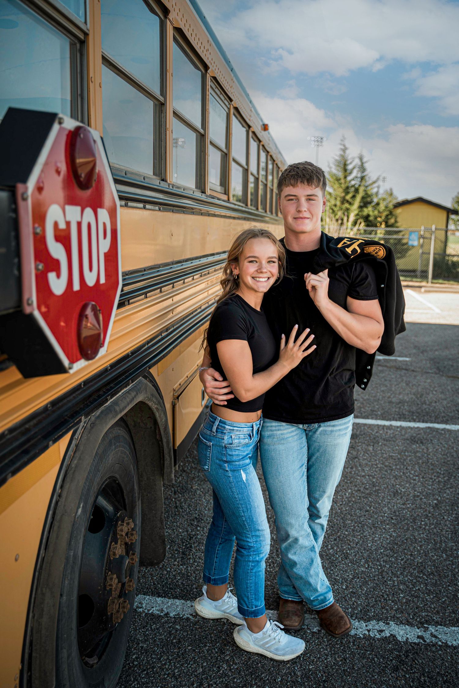 Two people in black shirts pose together beside a yellow school bus with a stop sign visible in the foreground.