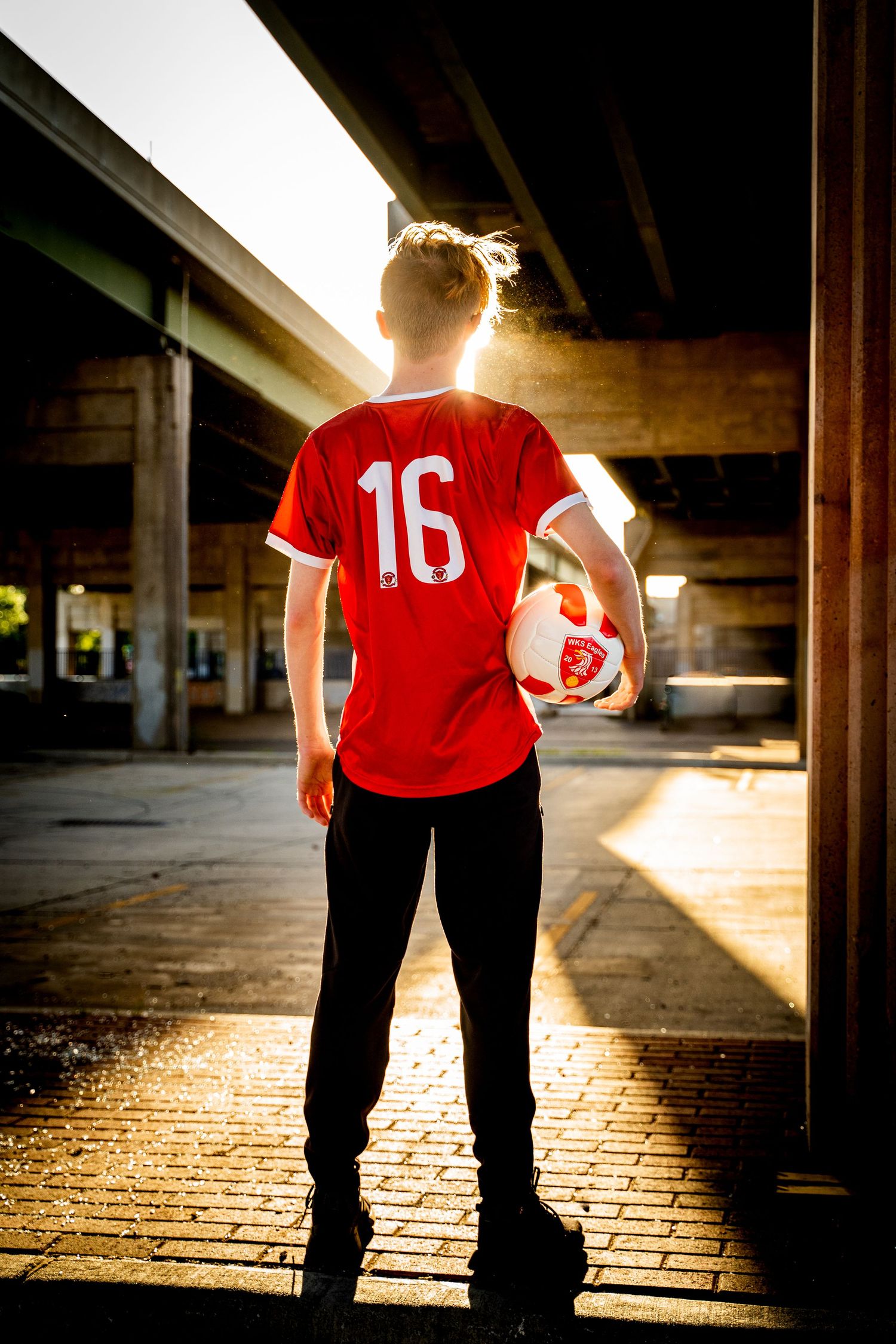 Athlete in red jersey number 16 stands silhouetted against sunlit urban backdrop.