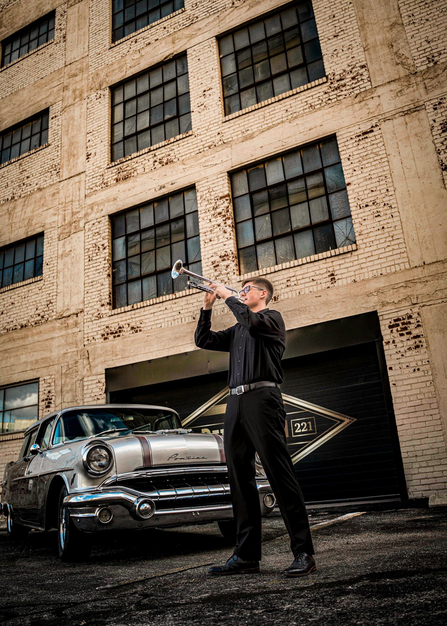 A vintage silver Pontiac parked next to an industrial brick building while someone drinks from a bottle in the foreground.