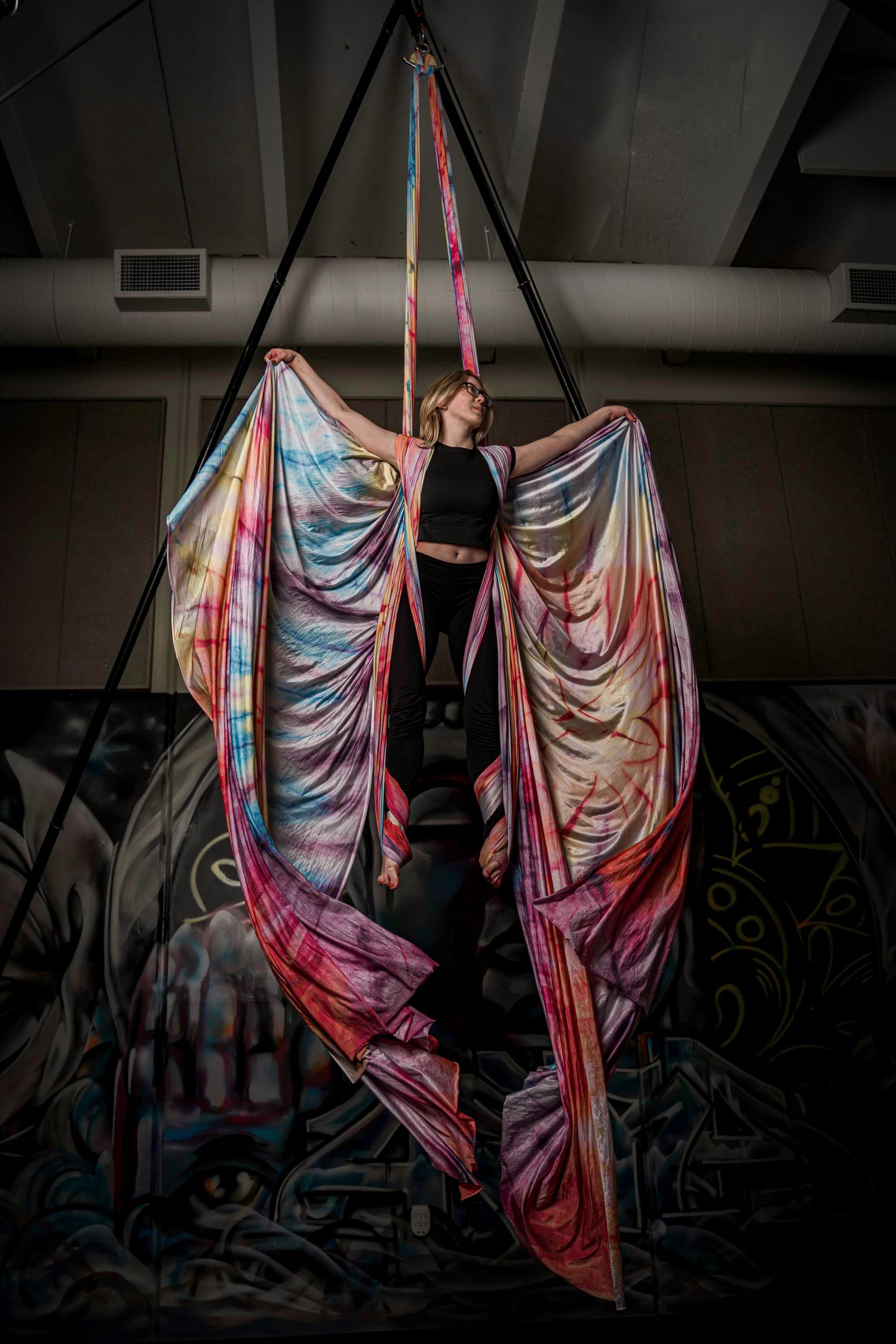 Aerial performer gracefully suspended in colorful silk fabric against dark theatrical backdrop during artistic performance.