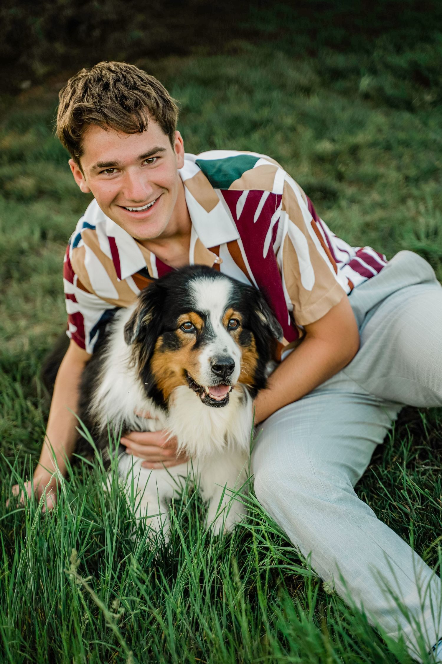 An Australian Shepherd dog with black and white fur sits in green grass with someone wearing a patterned shirt.