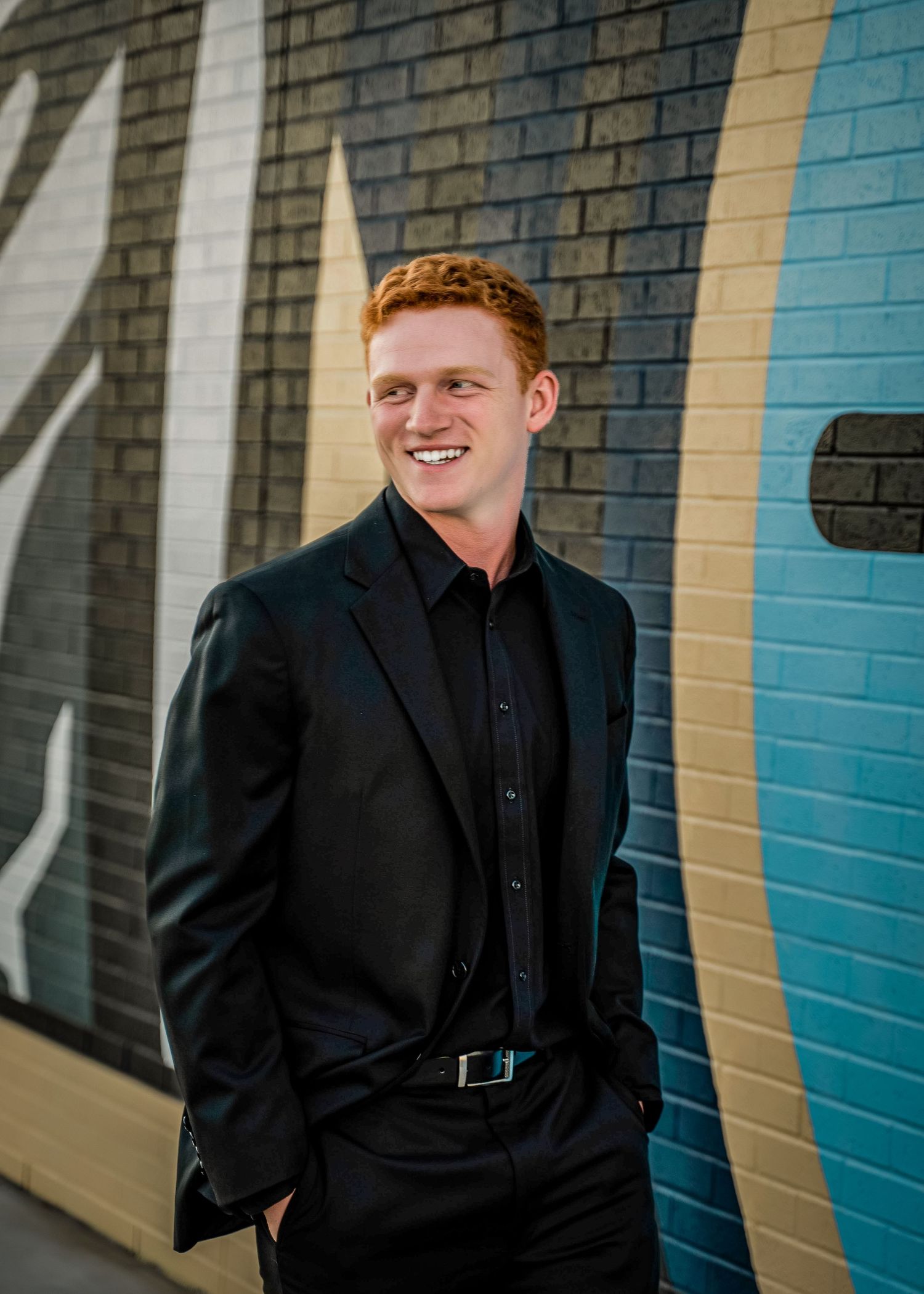 Professional portrait of a person in an all-black suit and shirt smiling against a colorful striped wall background.