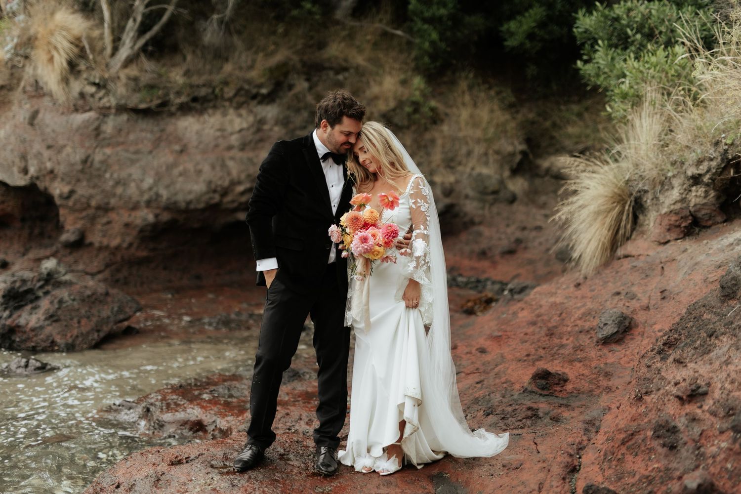 Wedding couple sharing an intimate moment on rocky terrain with coral-colored bouquet.