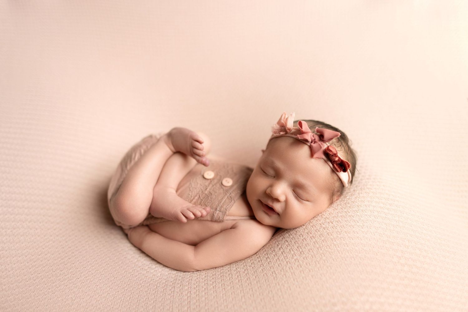 Sleeping newborn baby wearing floral headband and lace wrap on pink background.