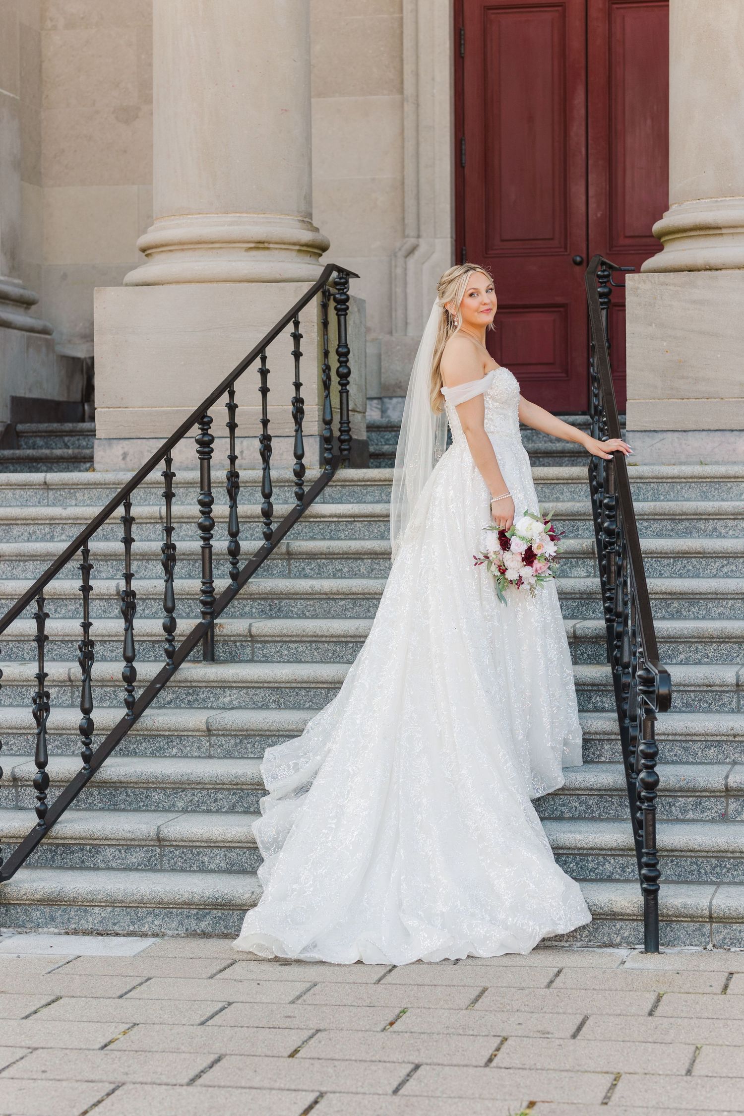 A bride in a flowing white gown poses on stone steps with ornate railings.