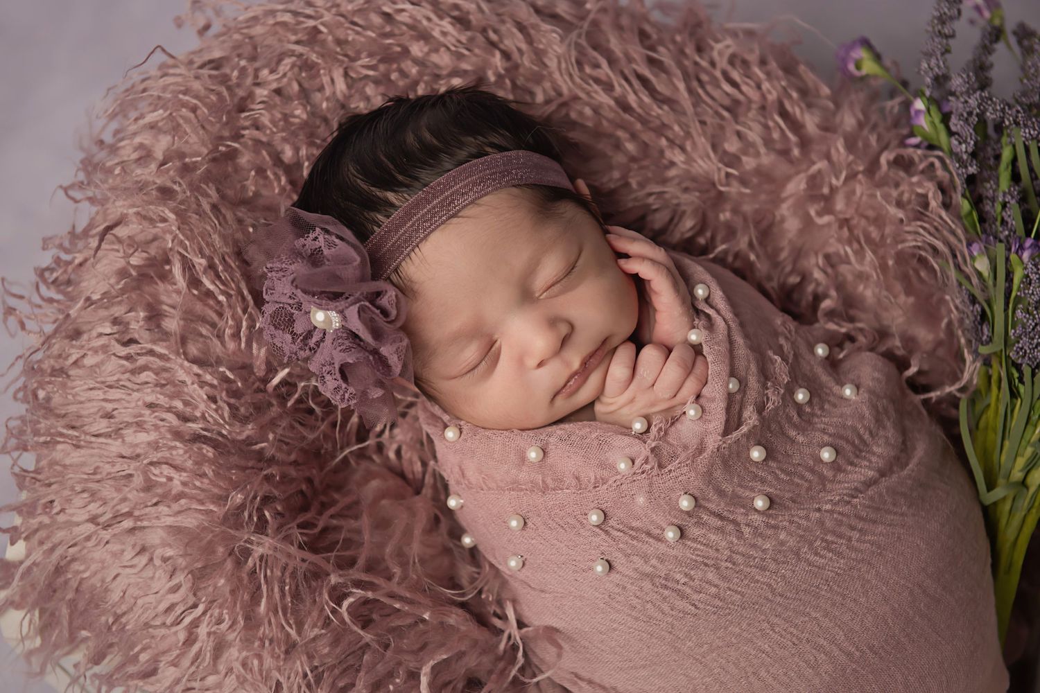 Newborn baby sleeping on pink textured blanket with lavender sprigs and pearl decorations.
