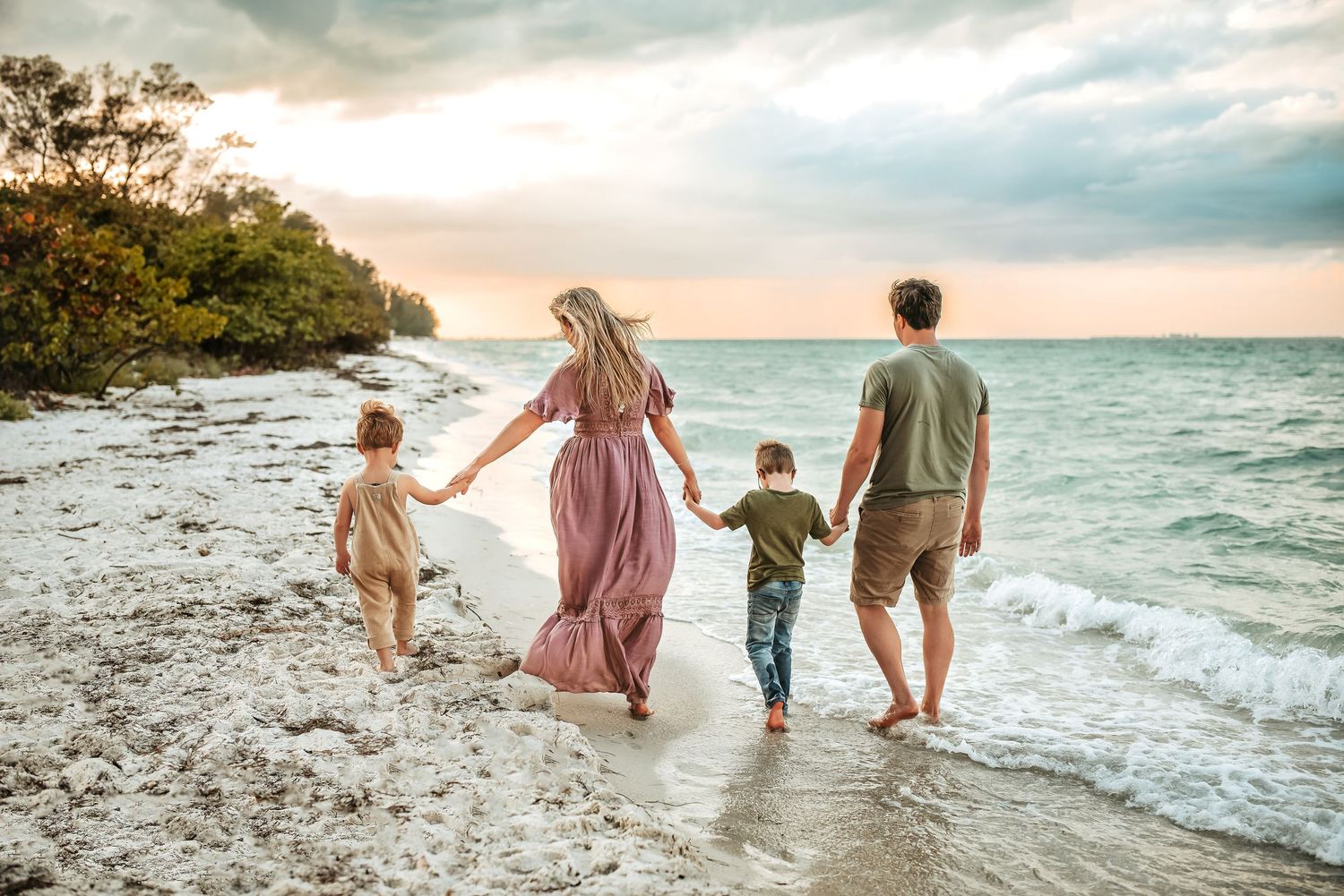 Family of four with two young boys walking down the beach together, holding hands, walking away.
