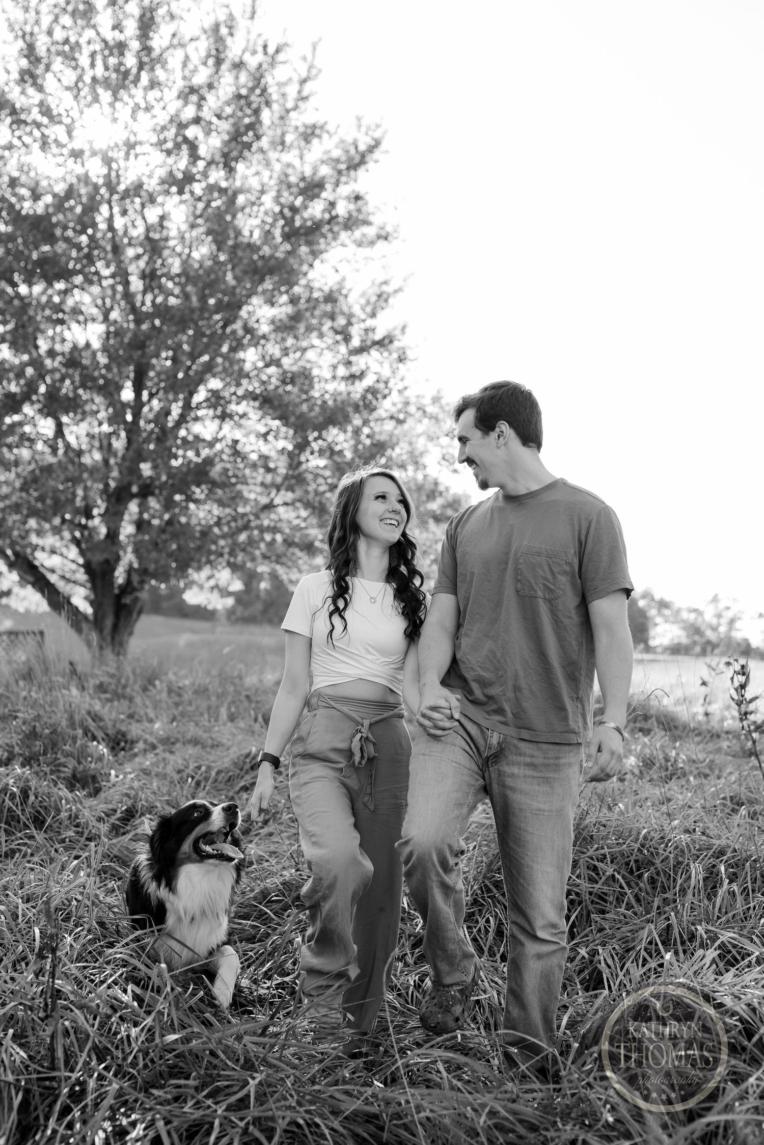 A black and white photo of a couple standing in tall grass with their Border Collie.
