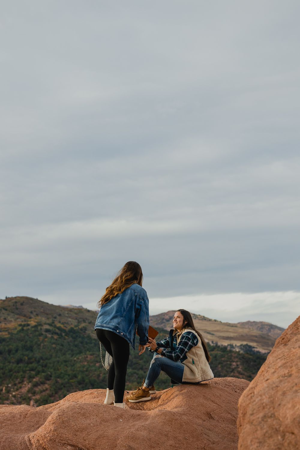 Two friends chatting while sitting on red rocks overlooking mountain vistas on a cloudy day.