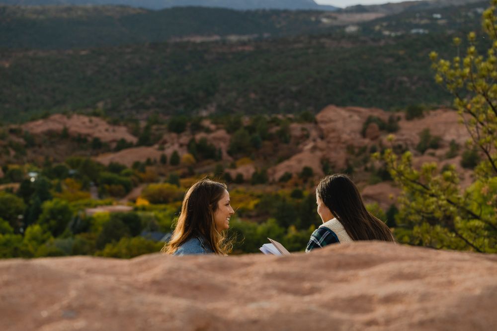 Two people sitting on red rocks enjoying a scenic mountain overlook with greenery below.
